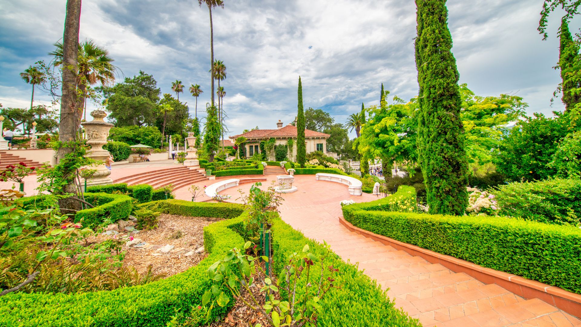 A grand estate with a large Mediterranean-style house and extensive terraced gardens featuring manicured hedges, statues, and tall trees under a partly cloudy sky.