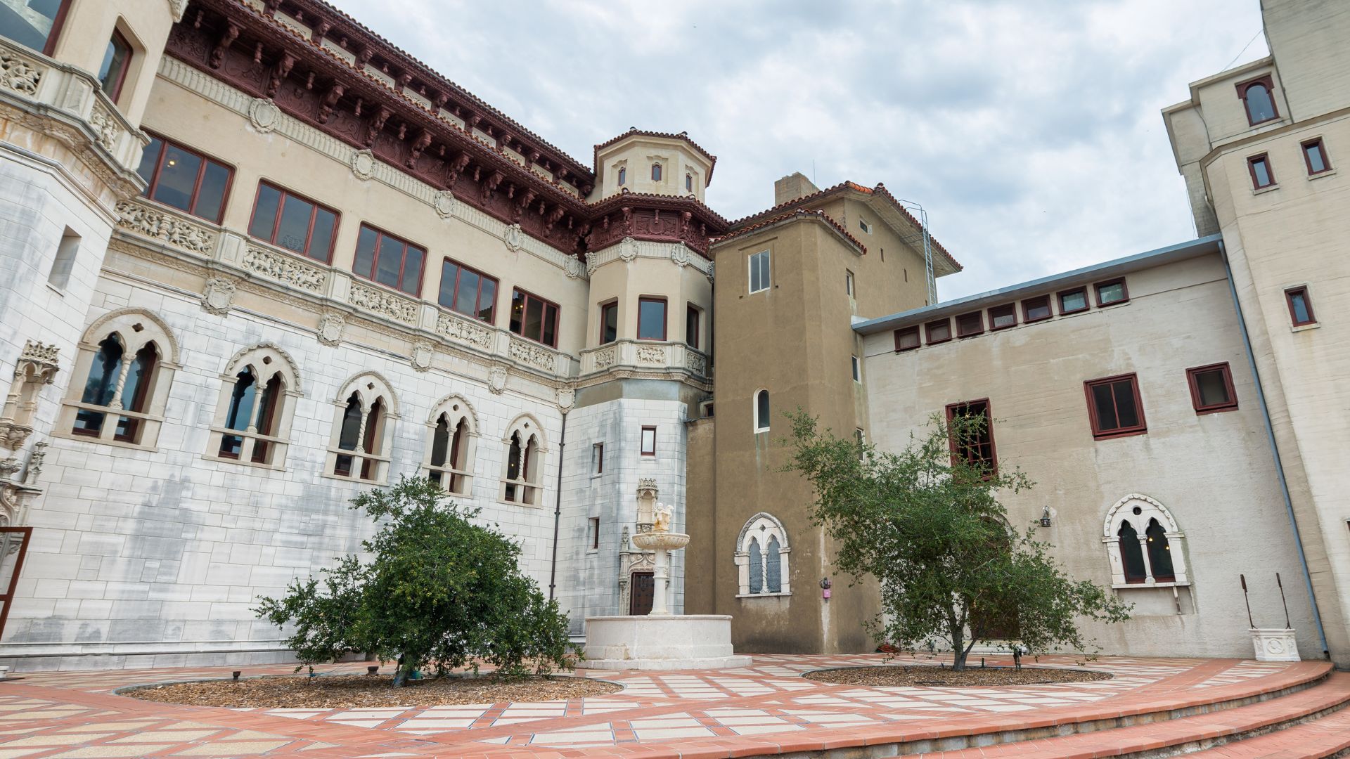 A grand, multi-storied building with ornate architectural details, including arched windows and balconies, surrounded by a paved courtyard with small trees, under a cloudy sky. The building features a mix of architectural styles, including elements resembling Gothic and Spanish Revival architecture.