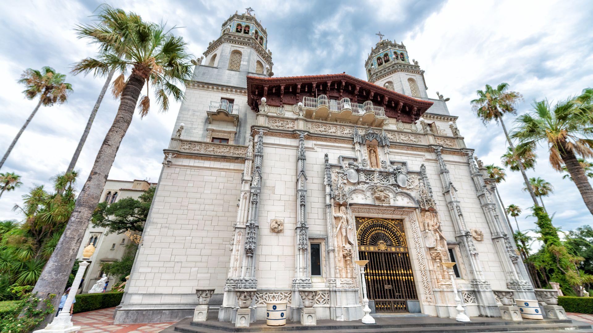 A low-angle exterior view of the grand, ornate, white stone facade of Hearst Castle, featuring prominent towers, intricate architectural details, and a large golden gate at the entrance, framed by tall palm trees against a dramatic, cloudy sky.