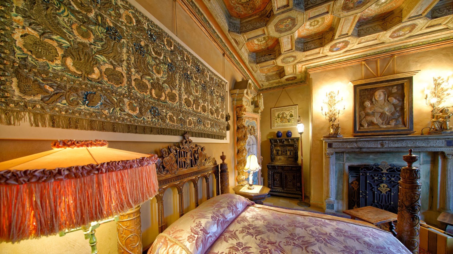 A richly decorated bedroom interior at Hearst Castle features a large, ornate bed with a patterned comforter and a fringed lampshade in the foreground. In the background, a grand fireplace with a decorative screen is visible, flanked by wall sconces and a framed painting above the mantle. The room boasts a highly detailed, patterned ceiling and a large, intricate tapestry covering a significant portion of one wall. 