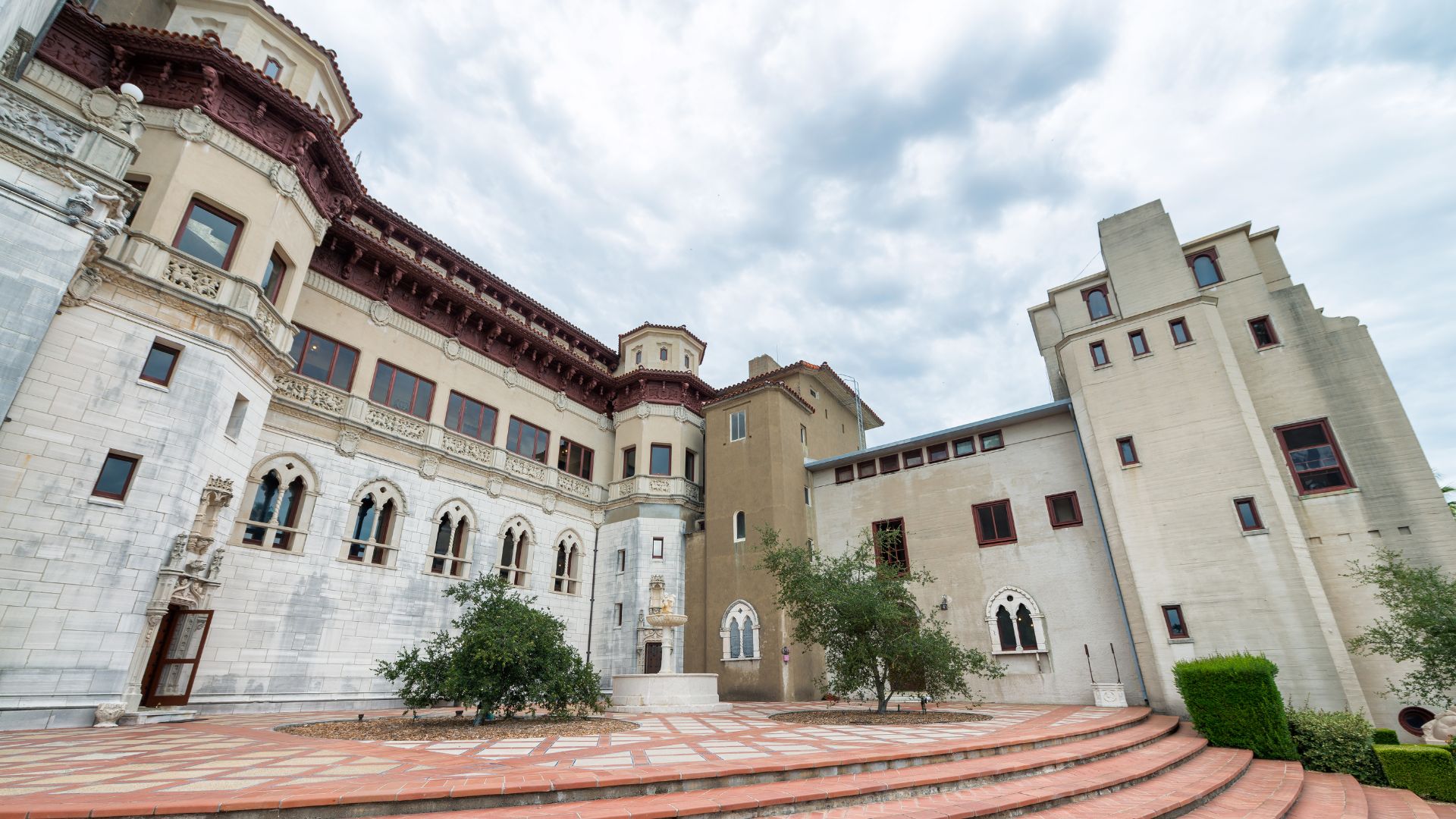 A wide-angle view of the exterior of Hearst Castle, showcasing its distinct architectural styles, including a white, ornate section with arched windows and a plainer, taller section, all under a cloudy sky. The foreground features a large, terraced patio with a circular design and scattered trees.
