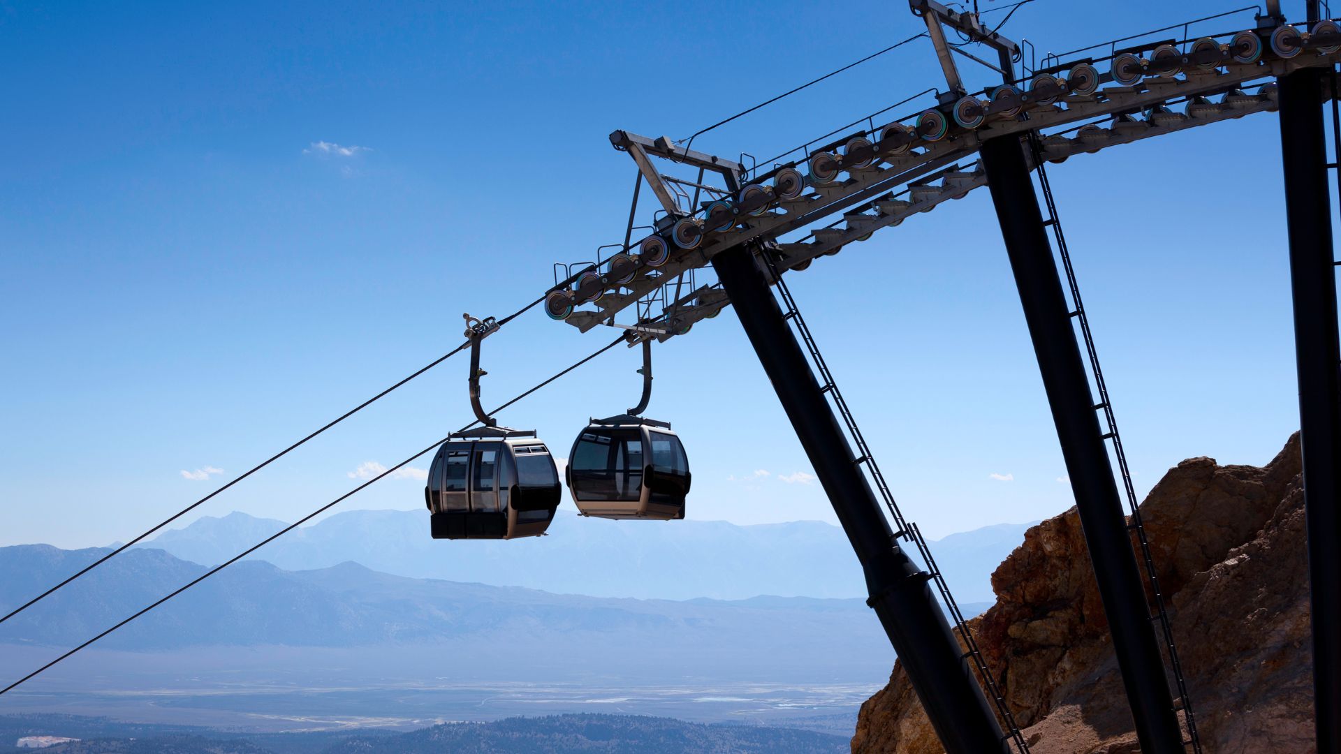 A vibrant image captures two gondola cars ascending a mountainside, with a vast mountain range stretching into the distance under a clear blue sky. The gondola support structure, made of dark metal, is prominently featured on the right, anchored to a rocky outcrop. The scene conveys a sense of elevation and expansive natural beauty.