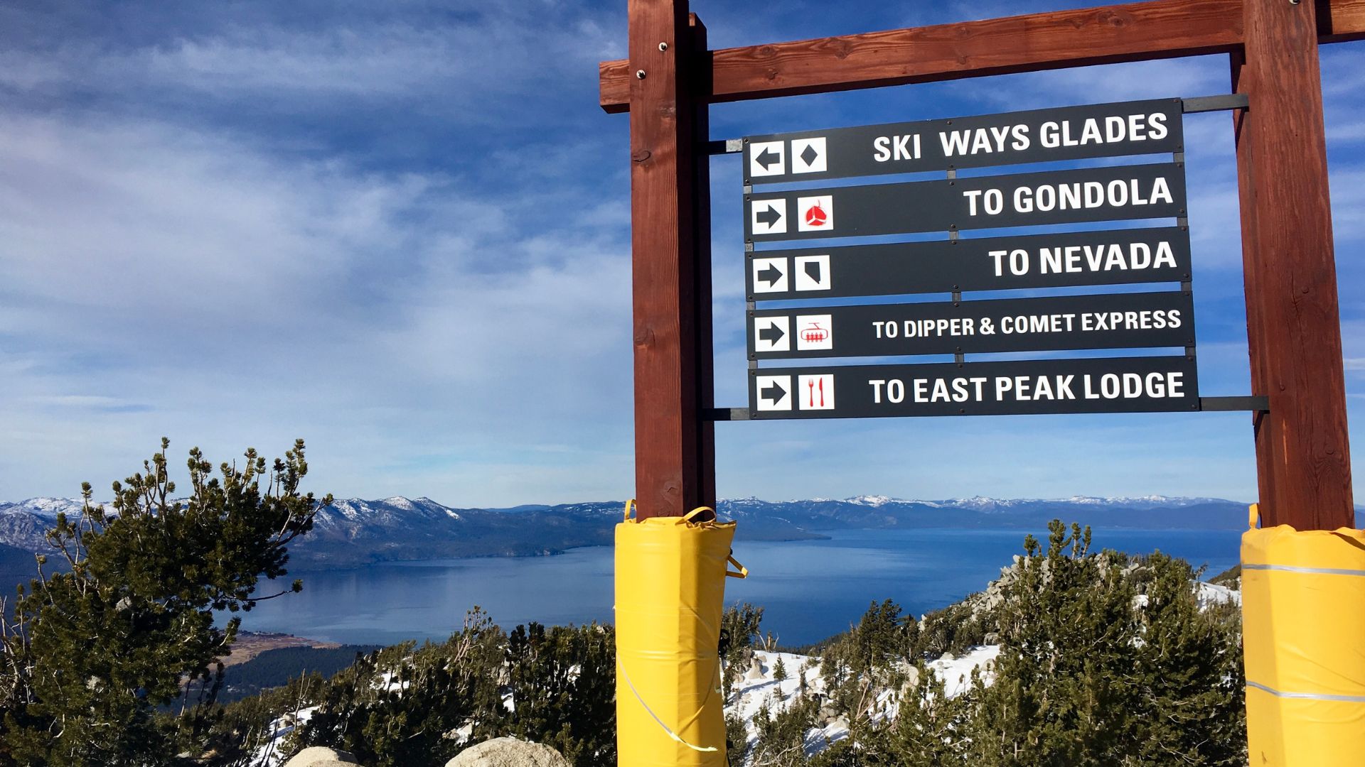 A wooden signpost at Heavenly Ski Resort in Lake Tahoe with directional signs pointing to ski runs and lodges, overlooking a panoramic view of Lake Tahoe and snow-covered mountains.