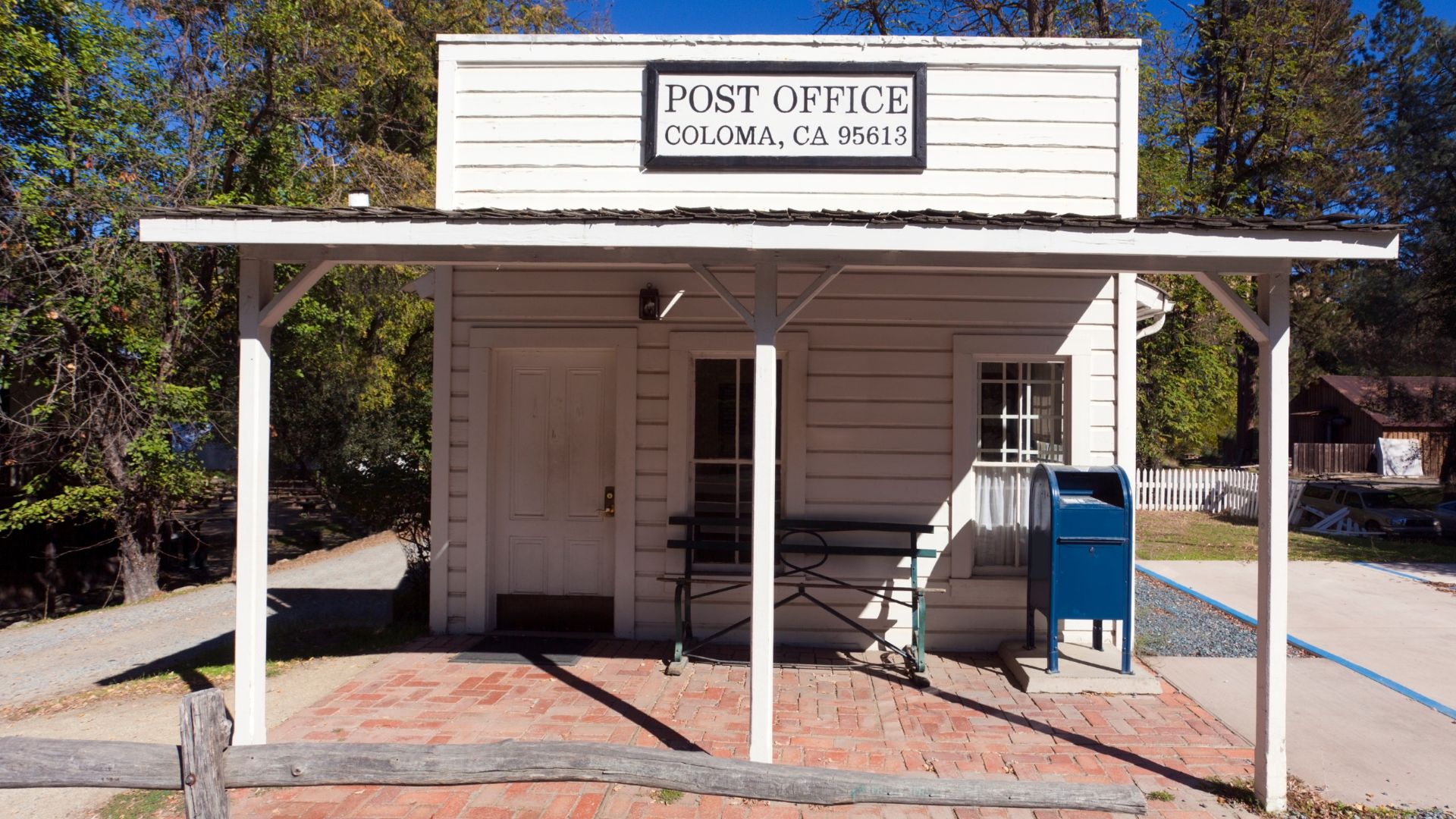 A small, white, wooden building with a sign that reads "POST OFFICE COLOMA, CA 95613" is visible under a white-framed porch supported by white pillars. A dark metal bench and a blue mailbox are situated on the brick patio in front of the building. Trees and a dirt path are visible in the background under a clear sky.