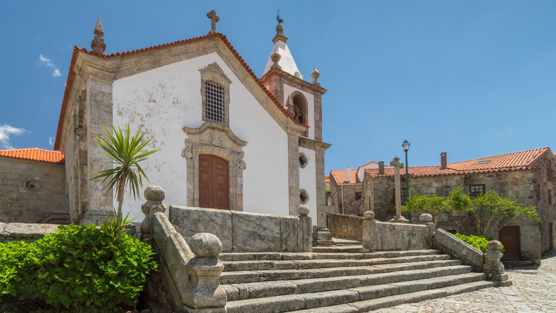 A historic white church with a prominent bell tower and a stone staircase leading up to its entrance, set against a clear blue sky in the village of Linhares da Beira, Central Portugal.