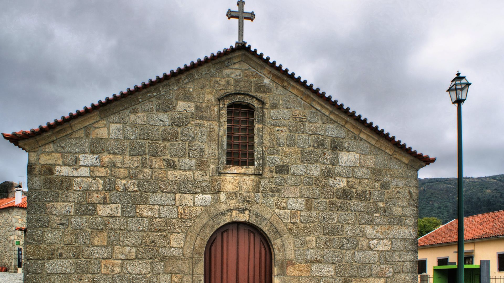 A stone church with a prominent cross on its roof and a rounded arch entrance, set against a cloudy sky, with a streetlamp and other buildings visible in the background.