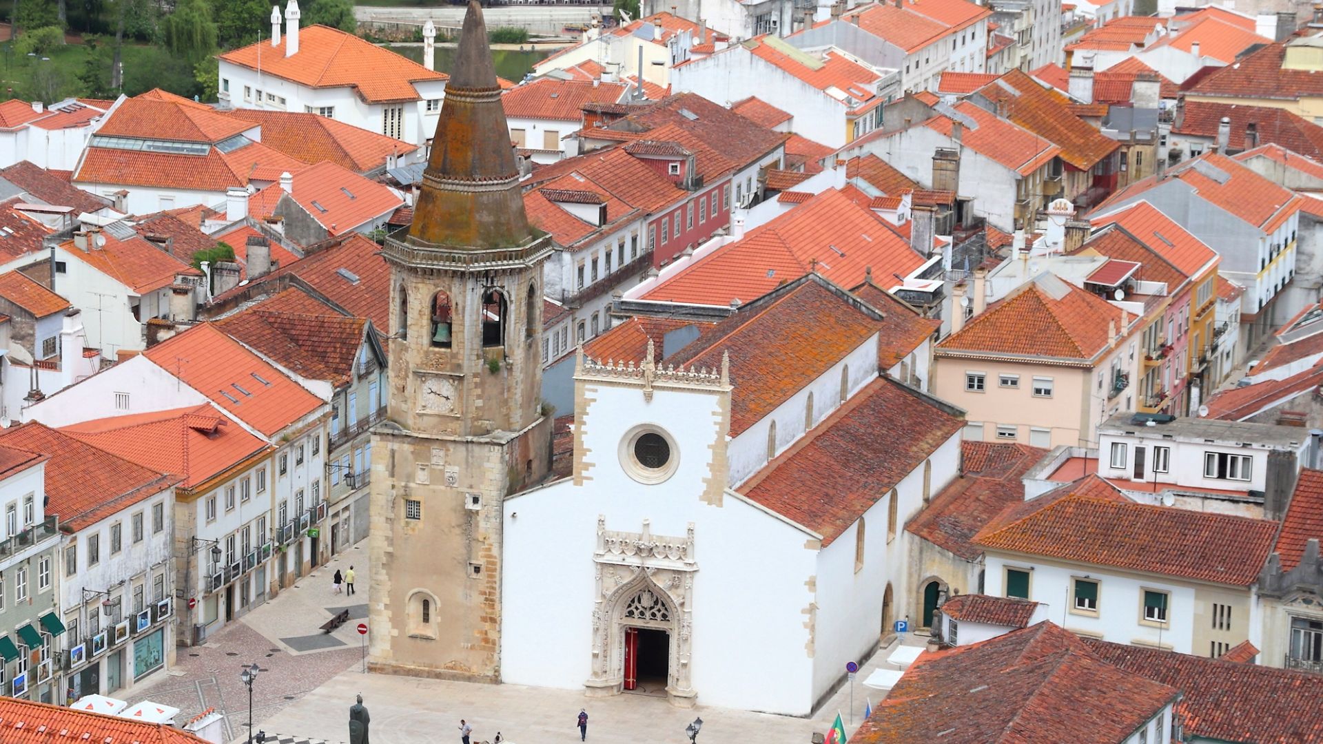 An aerial view captures the Igreja de São João Baptista in Tomar, Portugal, featuring its prominent bell tower and white facade, surrounded by a dense arrangement of terracotta-roofed buildings in the historic city center.