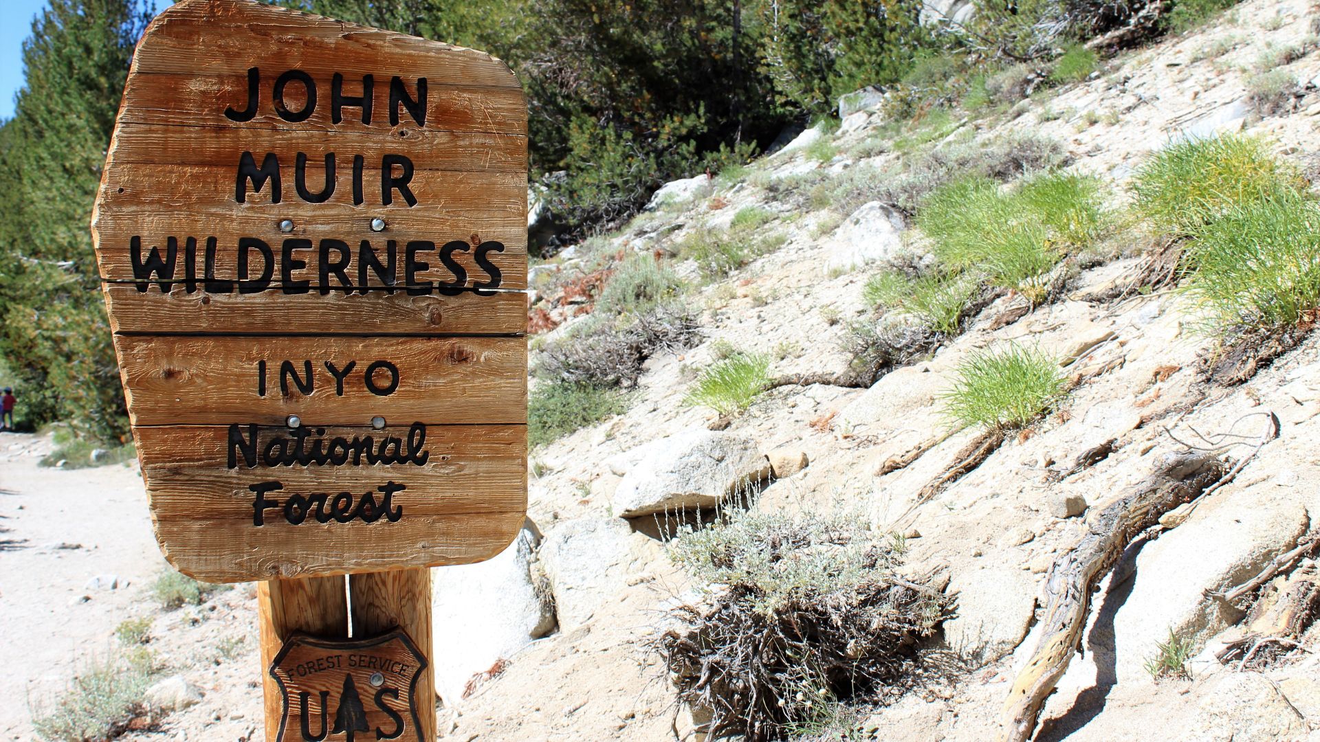 A weathered wooden sign stands on a rocky, brush-covered hillside, indicating "JOHN MUIR WILDERNESS" at the top and "INYO National Forest" below. The sign is set against a backdrop of a dirt path and pine trees in the distance.