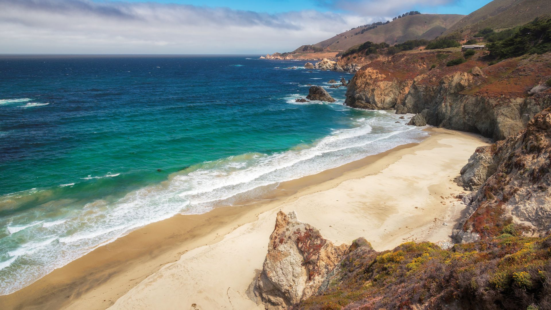 An aerial view of McWay Falls in Big Sur, California, showing an 80-foot waterfall cascading onto a sandy cove and flowing into the turquoise Pacific Ocean, framed by rugged cliffs and lush coastal vegetation under a clear sky.