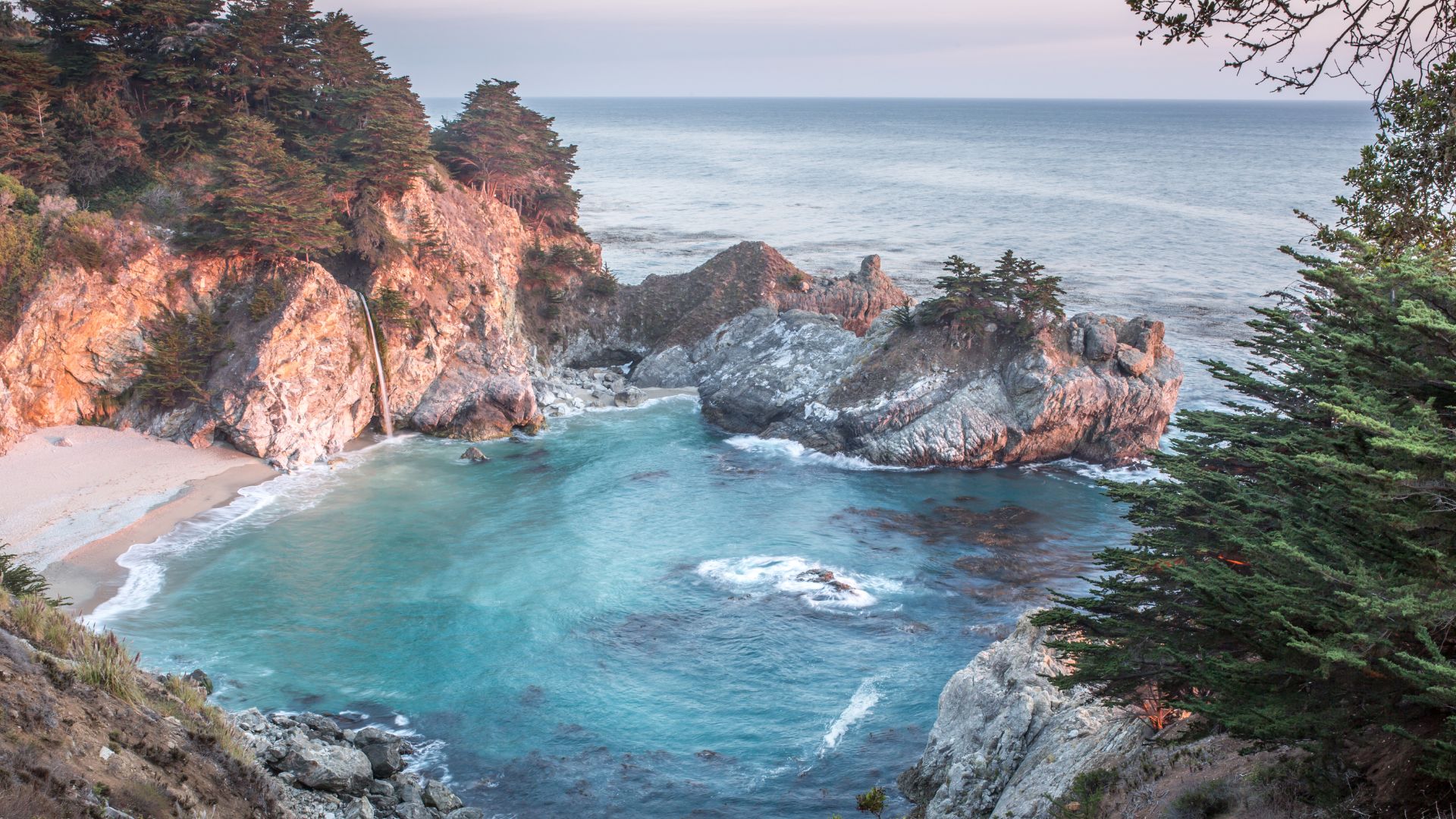 A scenic view of McWay Falls, an 80-foot waterfall cascading onto a sandy cove and into the turquoise waters of the Pacific Ocean, surrounded by rocky cliffs and lush green trees in Big Sur, California.