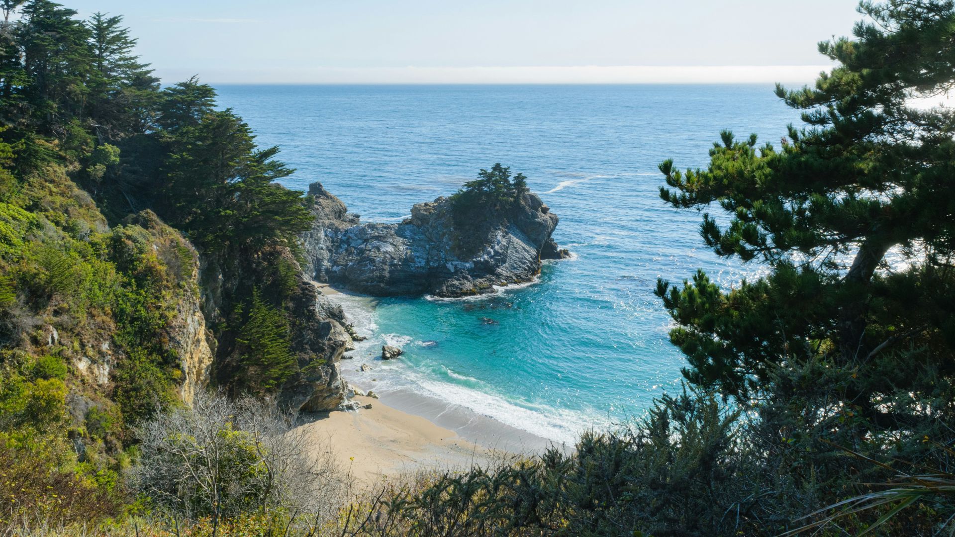 A scenic view of McWay Falls in Julia Pfeiffer Burns State Park, showing the 80-foot waterfall cascading onto a sandy beach in a picturesque cove, surrounded by lush green cliffs and the Pacific Ocean under a clear blue sky.