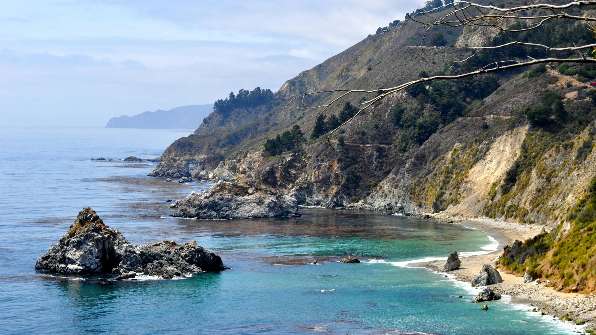 A scenic view of the California coastline at Julia Pfeiffer Burns State Park, featuring rugged cliffs, turquoise ocean waters, rocky outcrops, and a small secluded beach.