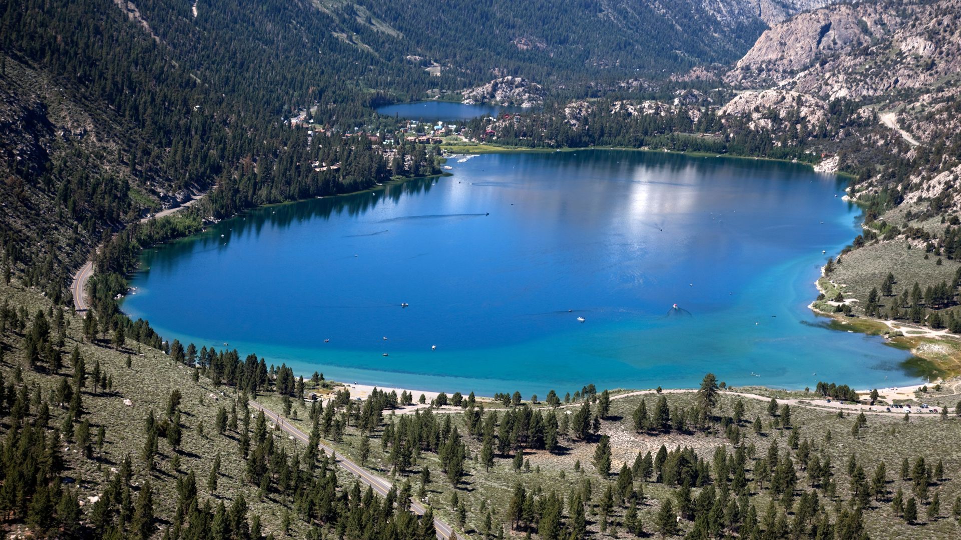 A long, winding highway stretches towards distant, snow-covered mountains under a clear sky, flanked by dry, brush-filled landscapes in the foreground.