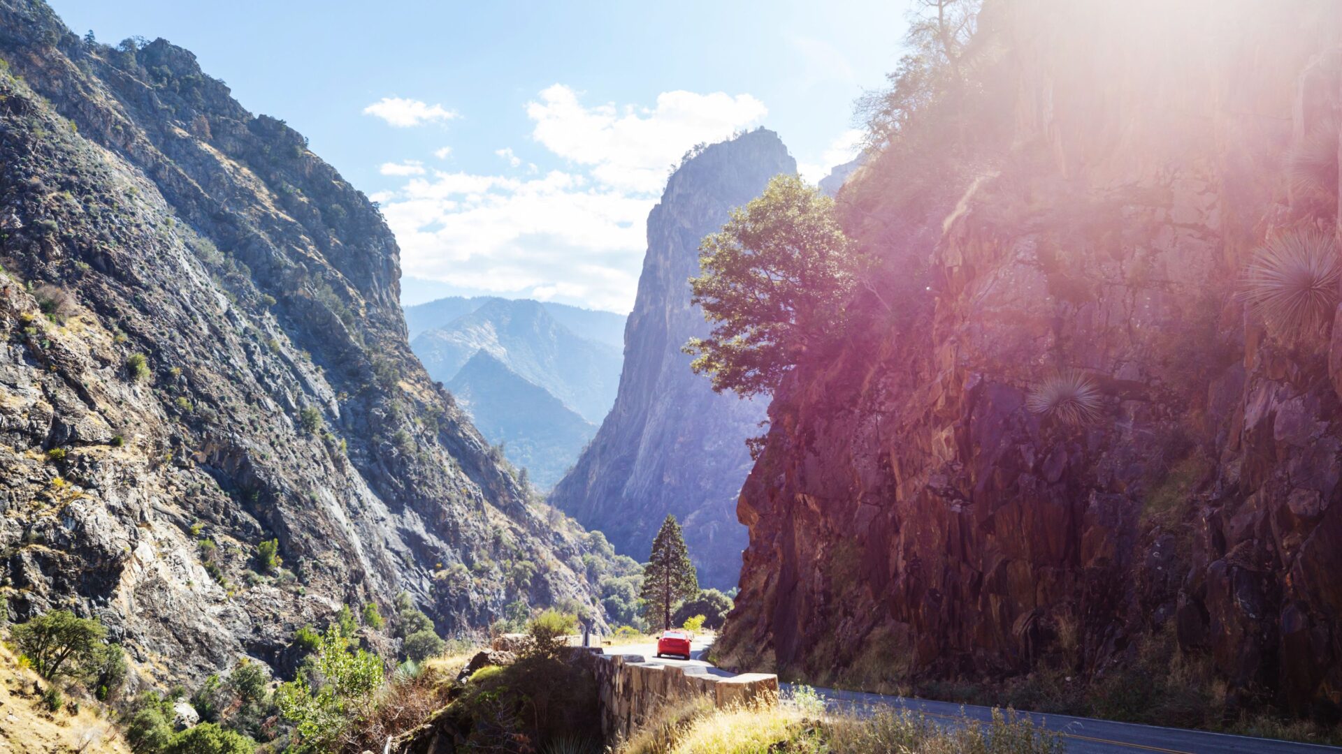 A wide shot of Kings Canyon National Park shows a deep canyon with steep, rocky cliffs on either side, a winding road at the bottom, and a small red vehicle parked on a viewpoint overlooking the valley. The scene is bathed in bright sunlight, highlighting the rugged terrain and distant mountains under a clear sky.