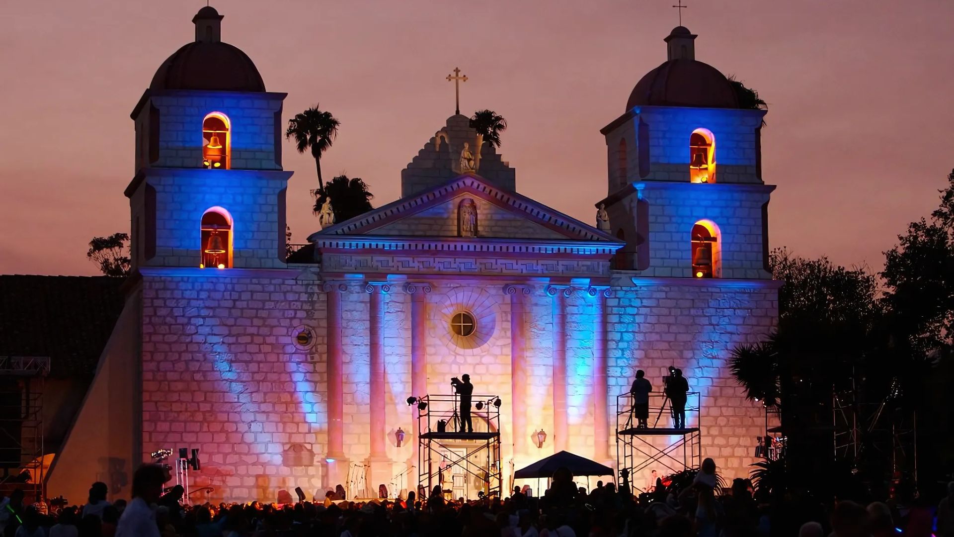 A vibrant twilight scene of the Old Mission Santa Barbara illuminated with blue and orange lights, with a crowd gathered in front and crew on scaffolding, during the La Fiesta Pequeña celebration.