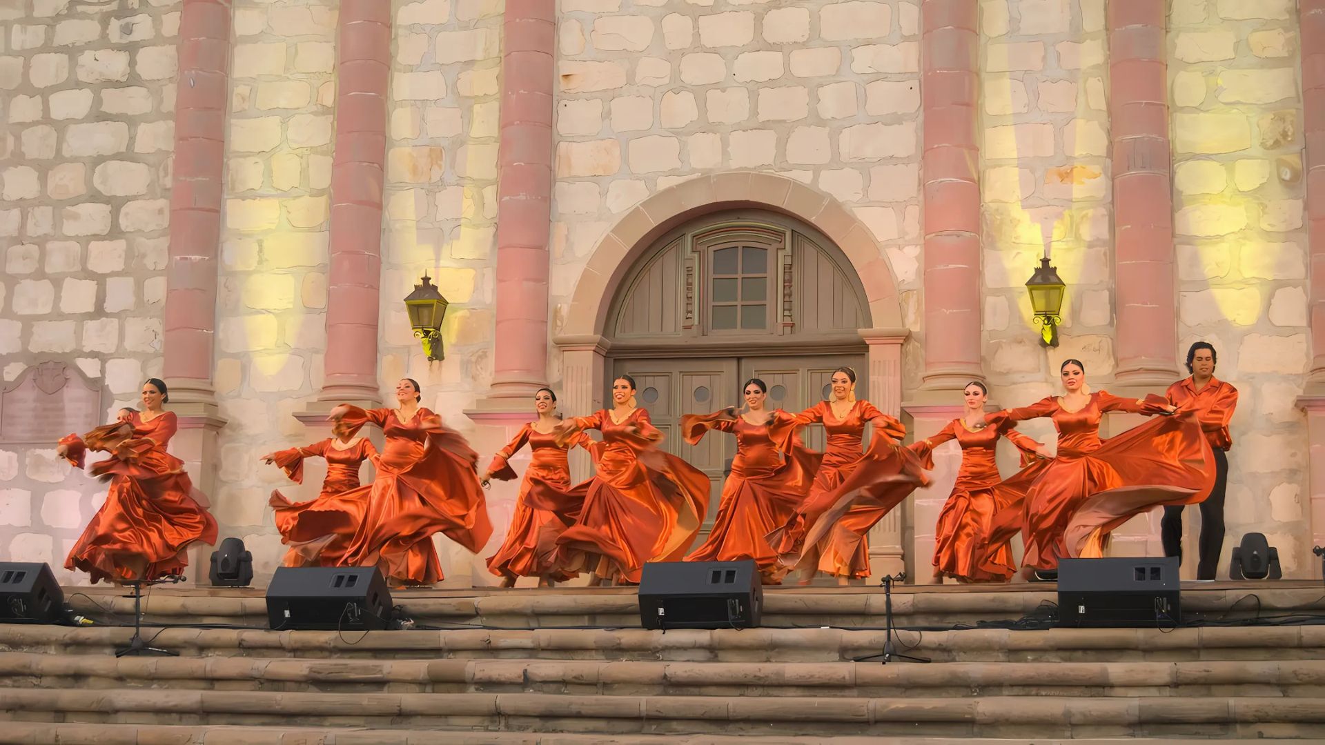 A group of performers in vibrant orange Spanish-style dresses dance on the steps of a stone building with arched doorways and pillars, illuminated by spotlights, during an evening event.