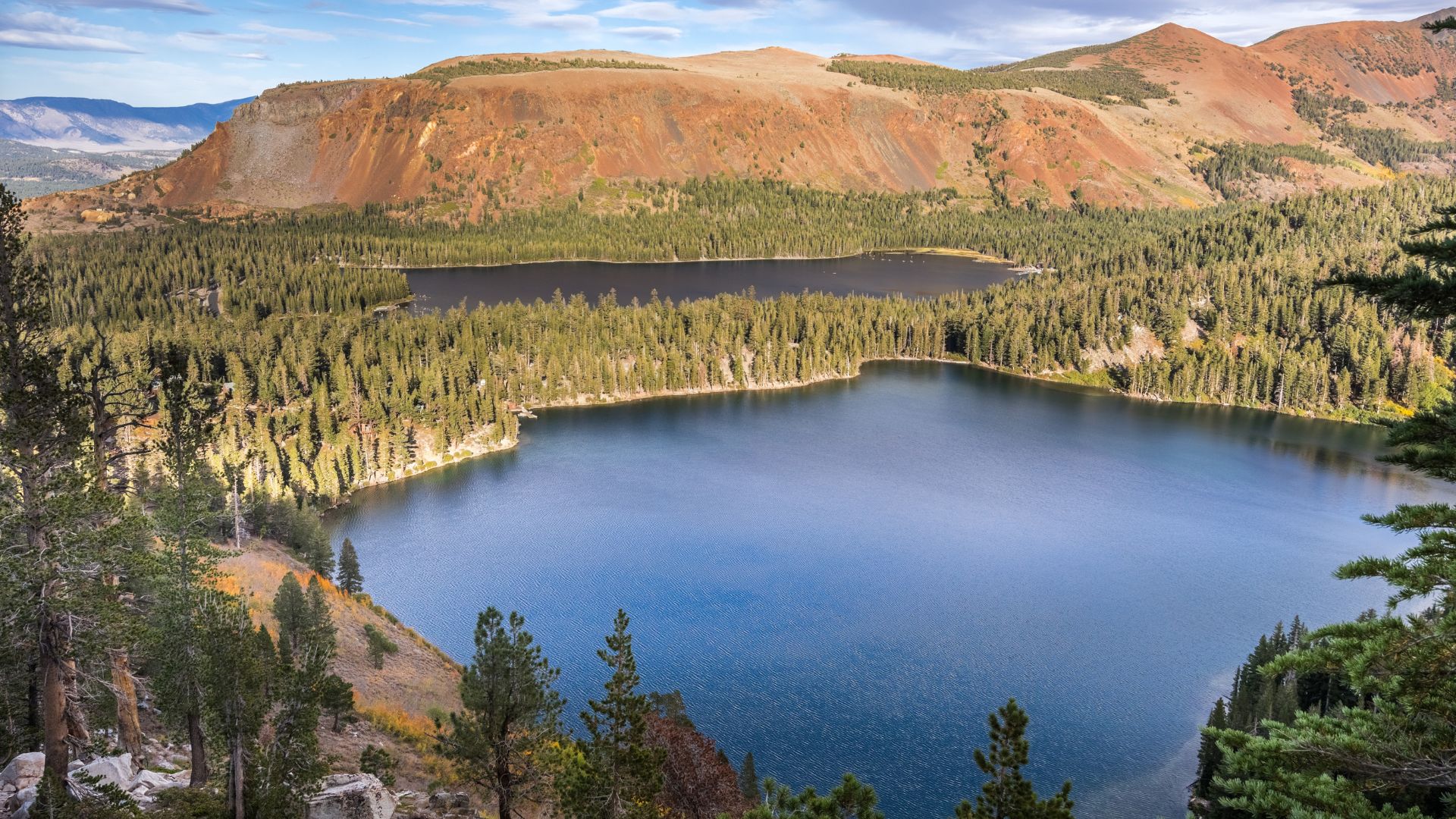 An aerial view of Lake George, a deep blue lake surrounded by dense evergreen forests and backed by reddish-brown mountains, with a smaller lake (Lake Marie) visible further in the distance under a clear sky.