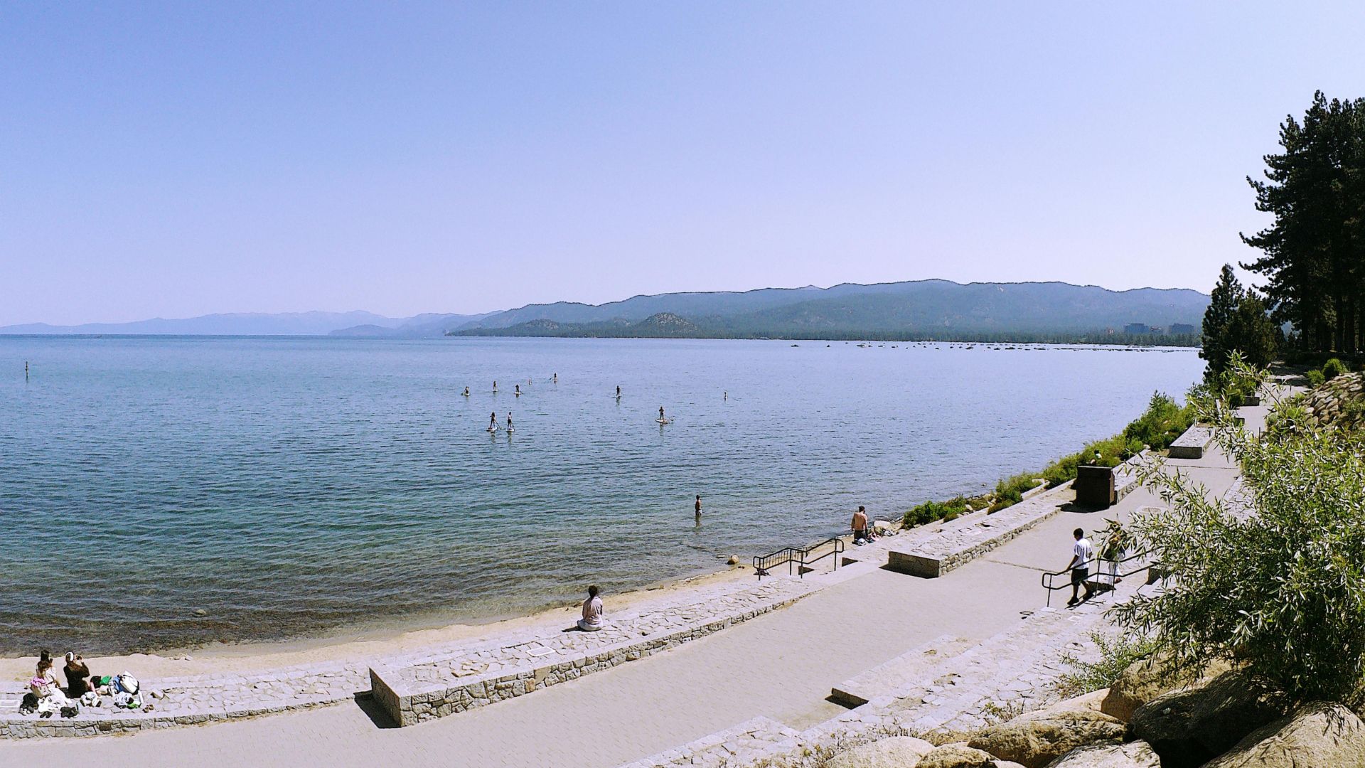 A wide-angle view of a calm Lake Tahoe under a clear blue sky, with a sandy beach and paved promenade in the foreground where people are relaxing and walking. Mountains are visible in the distance across the lake, and pine trees line the right side of the frame.