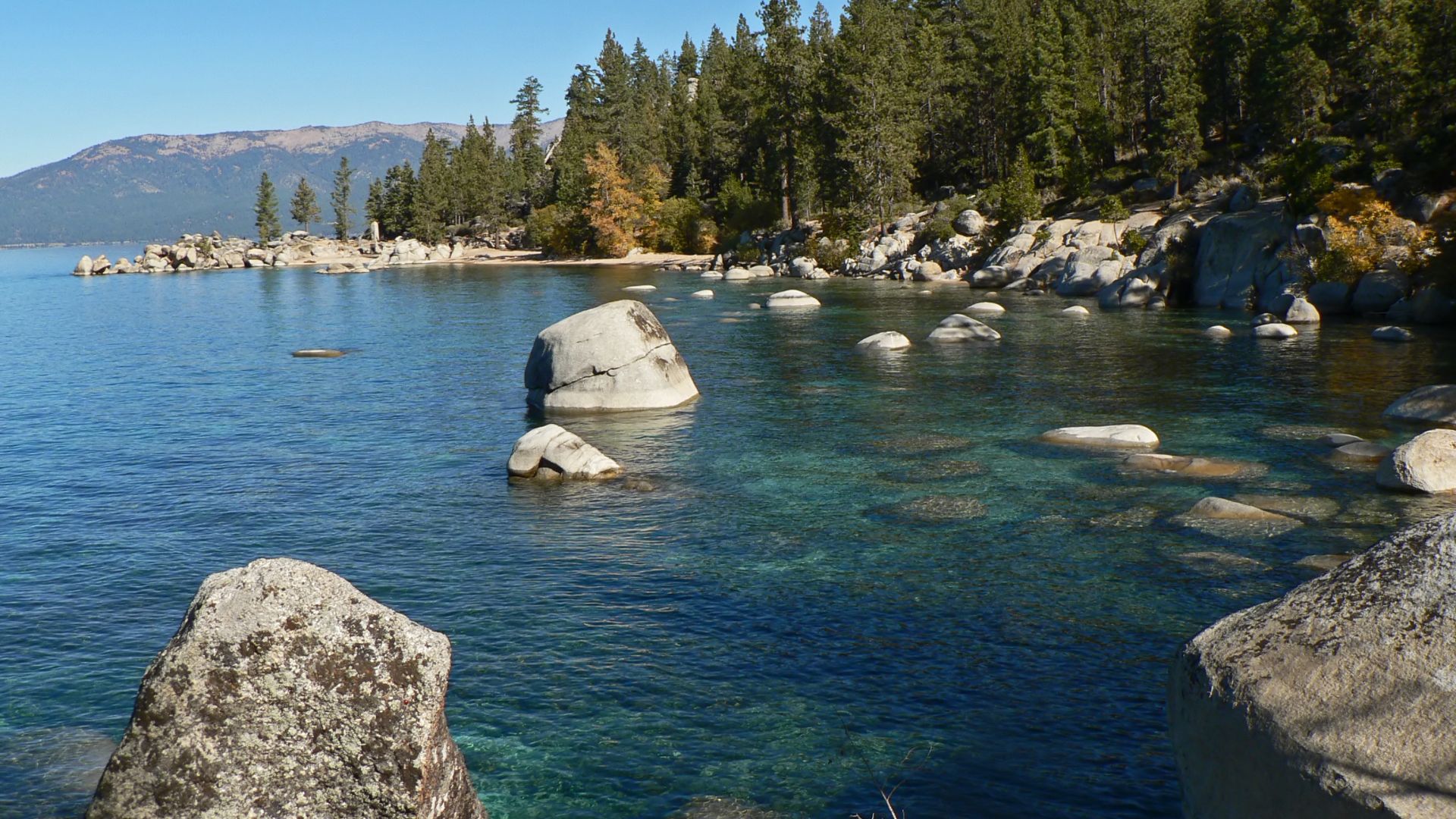 A clear, blue lake with large, smooth boulders in the foreground and extending into the water, surrounded by a shoreline with pine trees and distant mountains under a bright, clear sky.