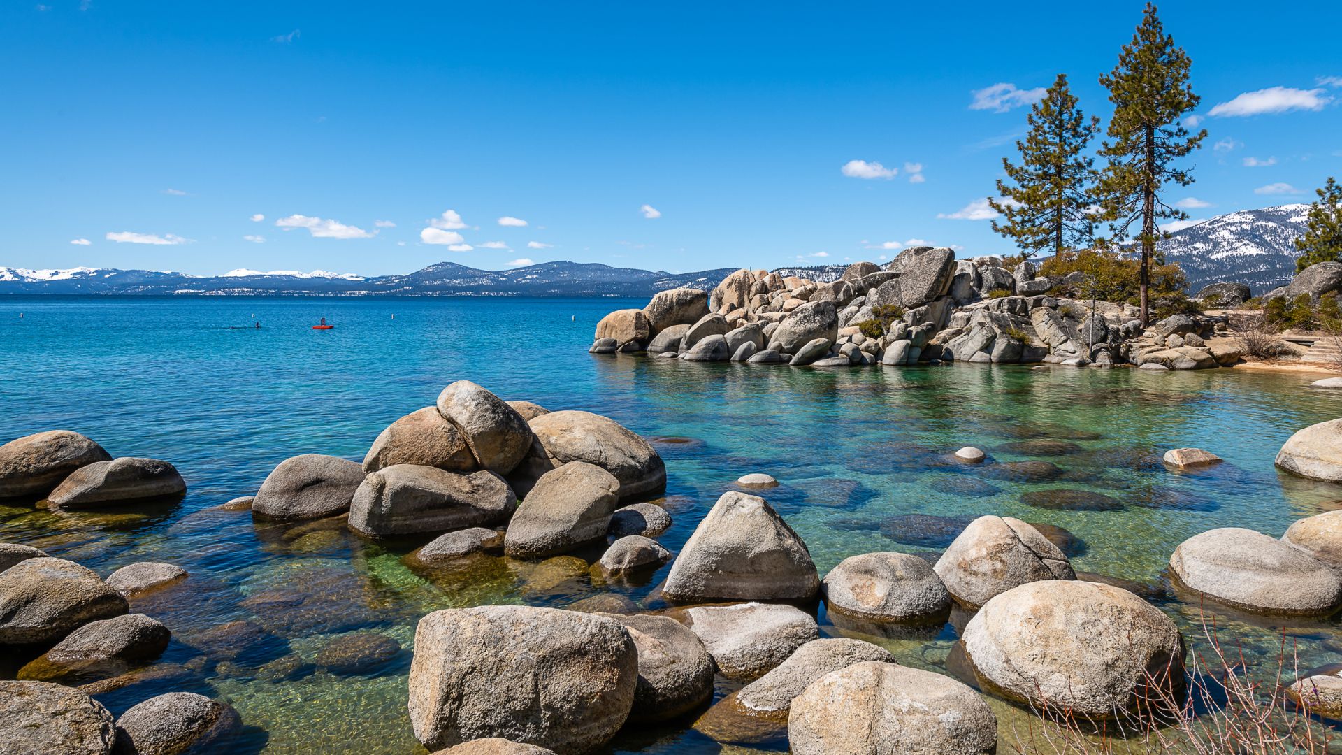 A scenic view of Lake Tahoe's clear blue water and rocky shoreline under a bright sky, with pine trees and snow-capped mountains in the background.