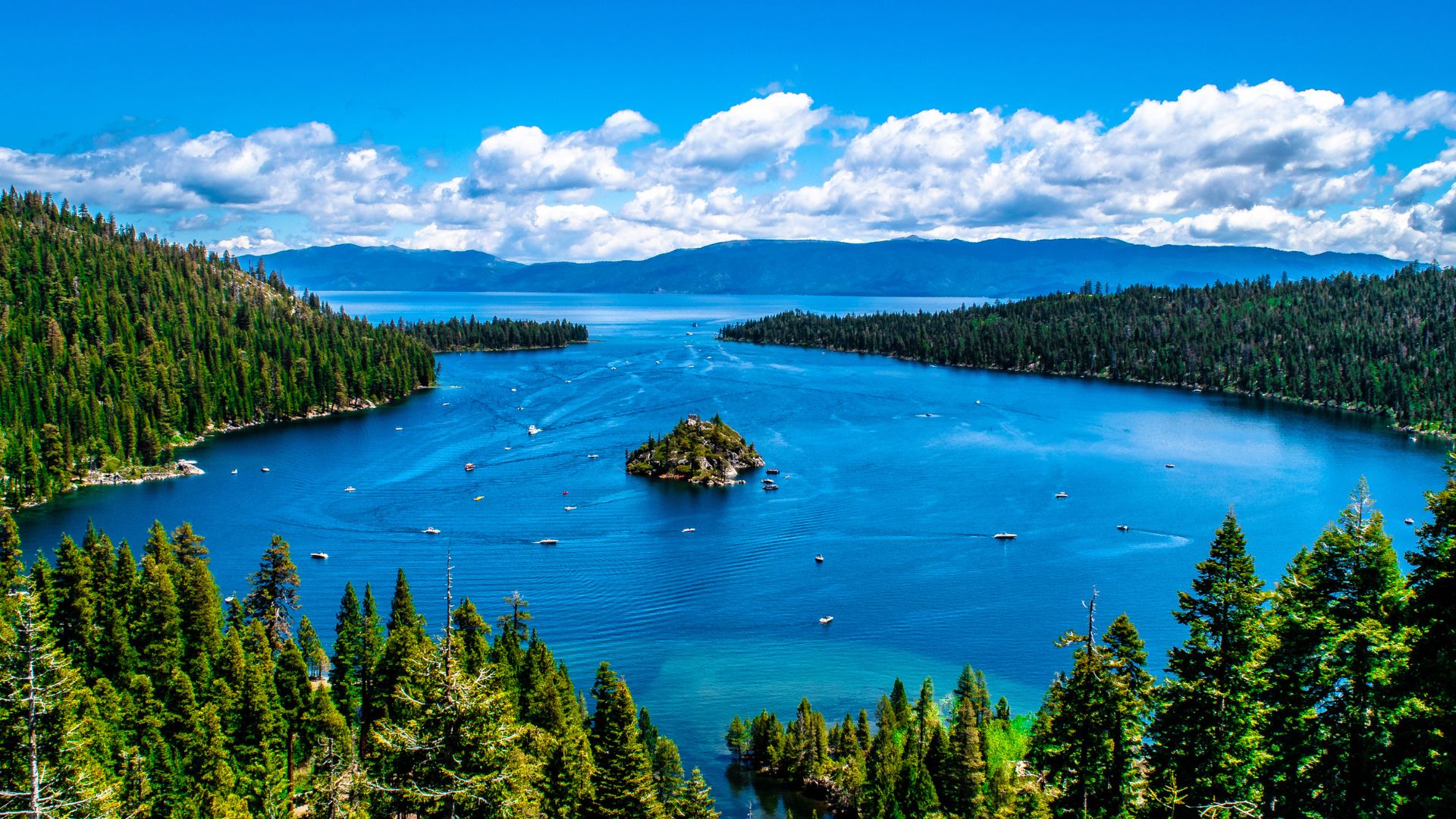 A wide shot of Lake Tahoe's clear blue waters, surrounded by dense green pine forests and distant mountains under a bright blue sky with scattered clouds. Several boats are visible on the lake, and a small, tree-covered island sits prominently in the center of the bay.