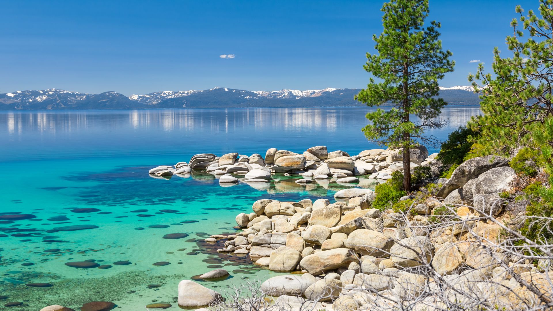 A vibrant image showcasing the clear, turquoise waters of Lake Tahoe, with a rocky shoreline in the foreground and snow-capped mountains visible in the distance under a clear blue sky.