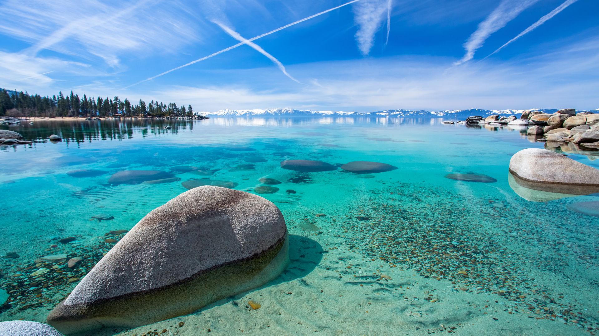 A vibrant image of Lake Tahoe featuring crystal-clear turquoise water with visible rocks on the lakebed in the foreground, surrounded by a forested shoreline and snow-capped mountains under a bright blue sky with prominent contrails.
