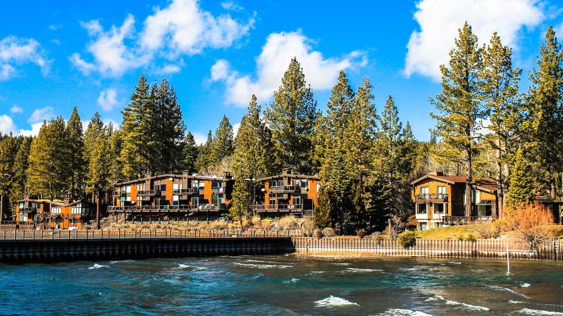 A panoramic view of a resort or lodging complex with multi-story buildings and balconies, nestled among tall evergreen trees along the edge of a wide river or lake under a blue sky with scattered clouds.