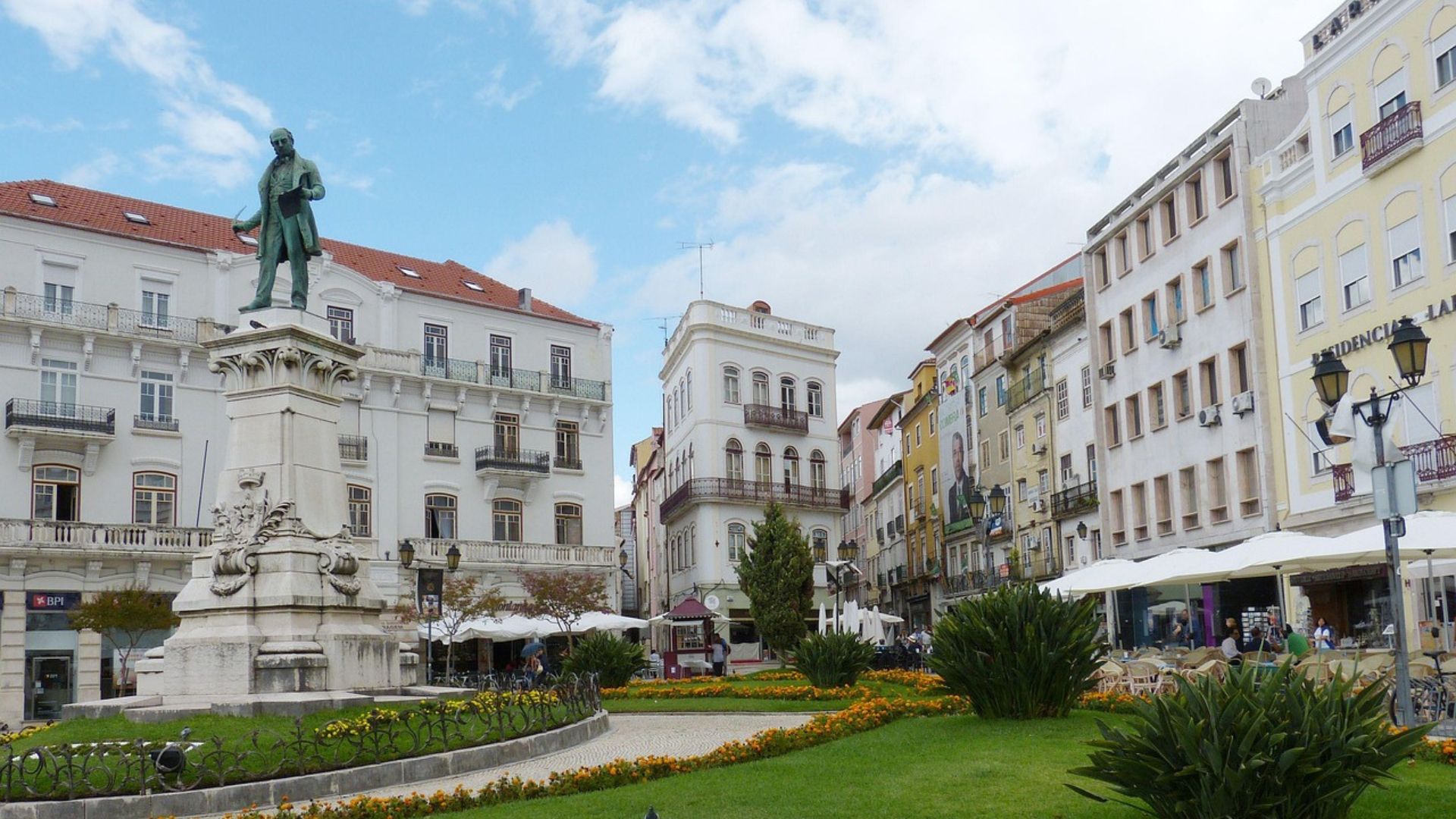 The image shows Largo da Portagem, a prominent square in Coimbra, Portugal, featuring a statue, historic buildings, and outdoor seating areas, indicating a lively urban setting. 