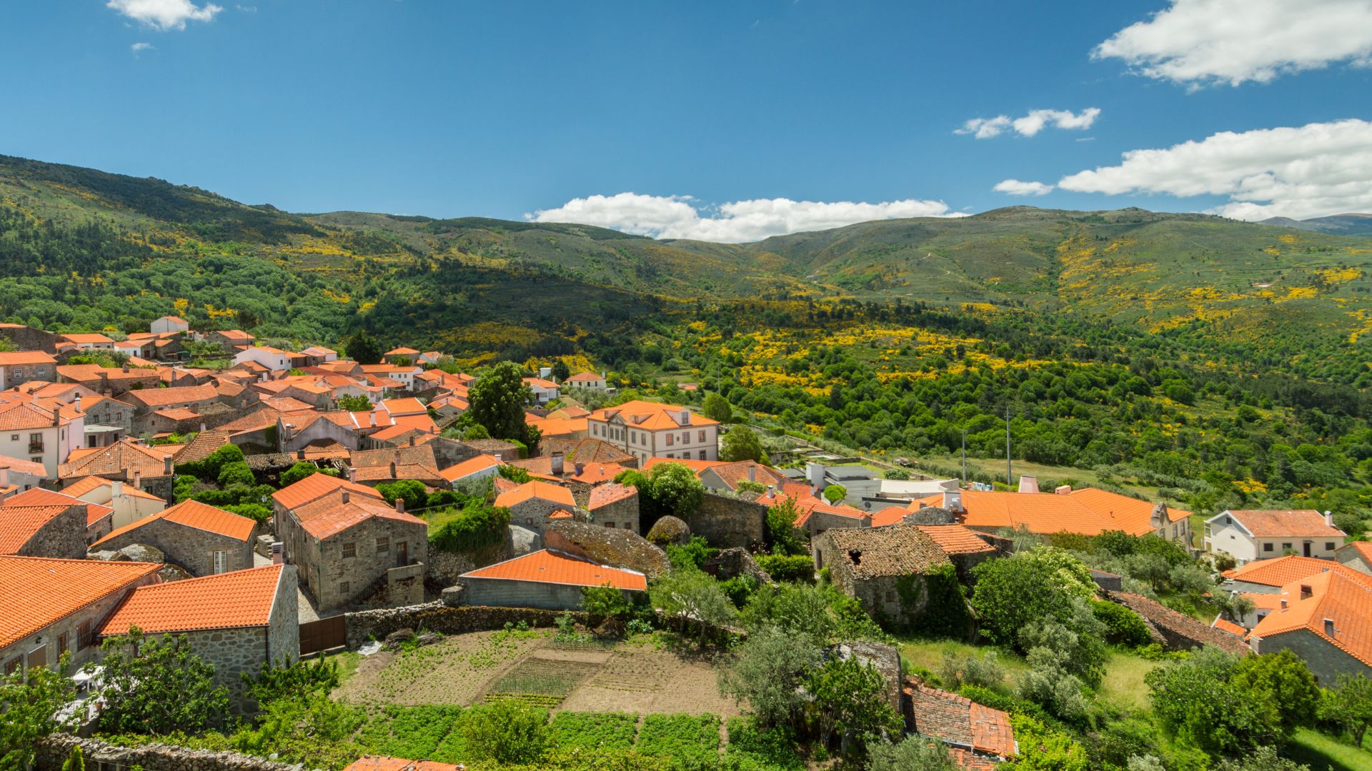 A panoramic view of Linhares da Beira, a historical village in Central Portugal, featuring traditional stone houses with orange tiled roofs nestled amidst lush green hills and mountains under a bright blue sky.