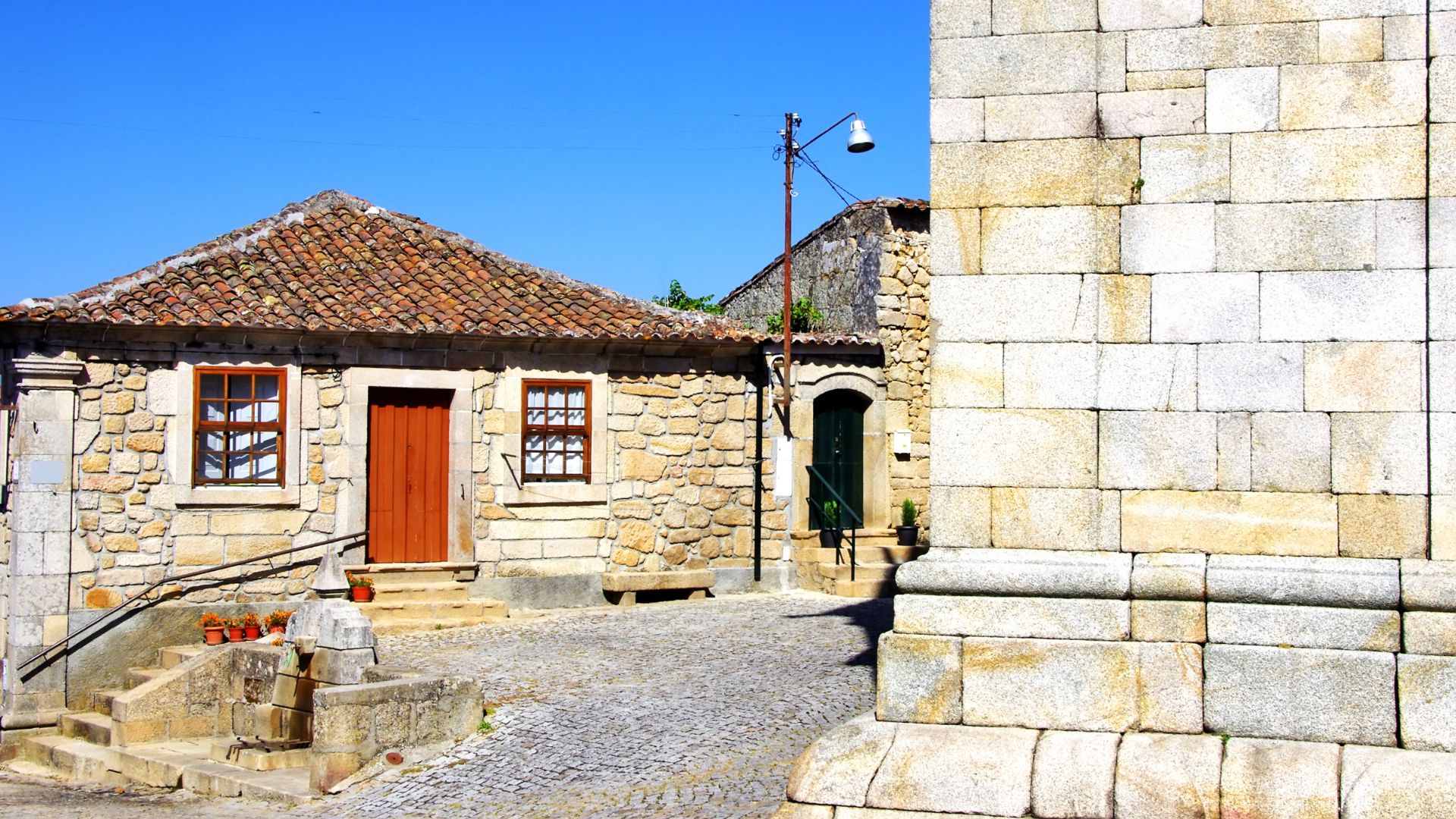 A narrow, sunlit cobblestone street in a historic Portuguese village, featuring traditional stone houses with red-tiled roofs and a prominent stone building on the right.