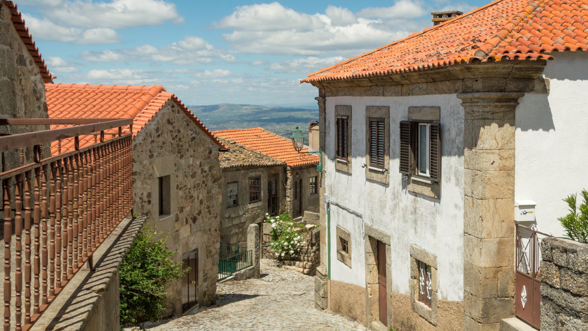 A narrow, cobblestone alleyway in a historic Portuguese village, featuring traditional stone houses with orange-tiled roofs under a partly cloudy sky.