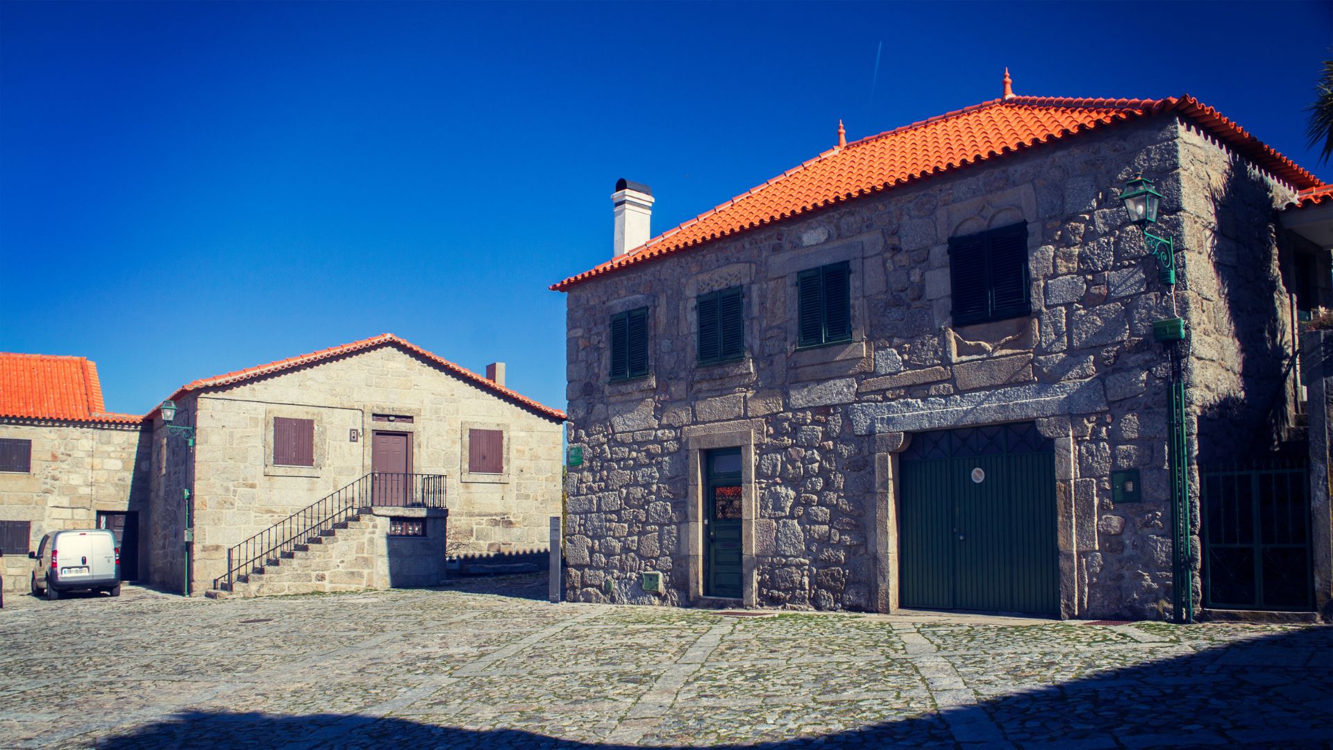 The image shows a cobblestone square in Linhares da Beira, Portugal, featuring traditional stone houses with red-tiled roofs under a clear blue sky. One house on the left has an external staircase leading to its entrance, while a larger building on the right features a prominent green wooden door. A white van is parked on the left side of the square. 