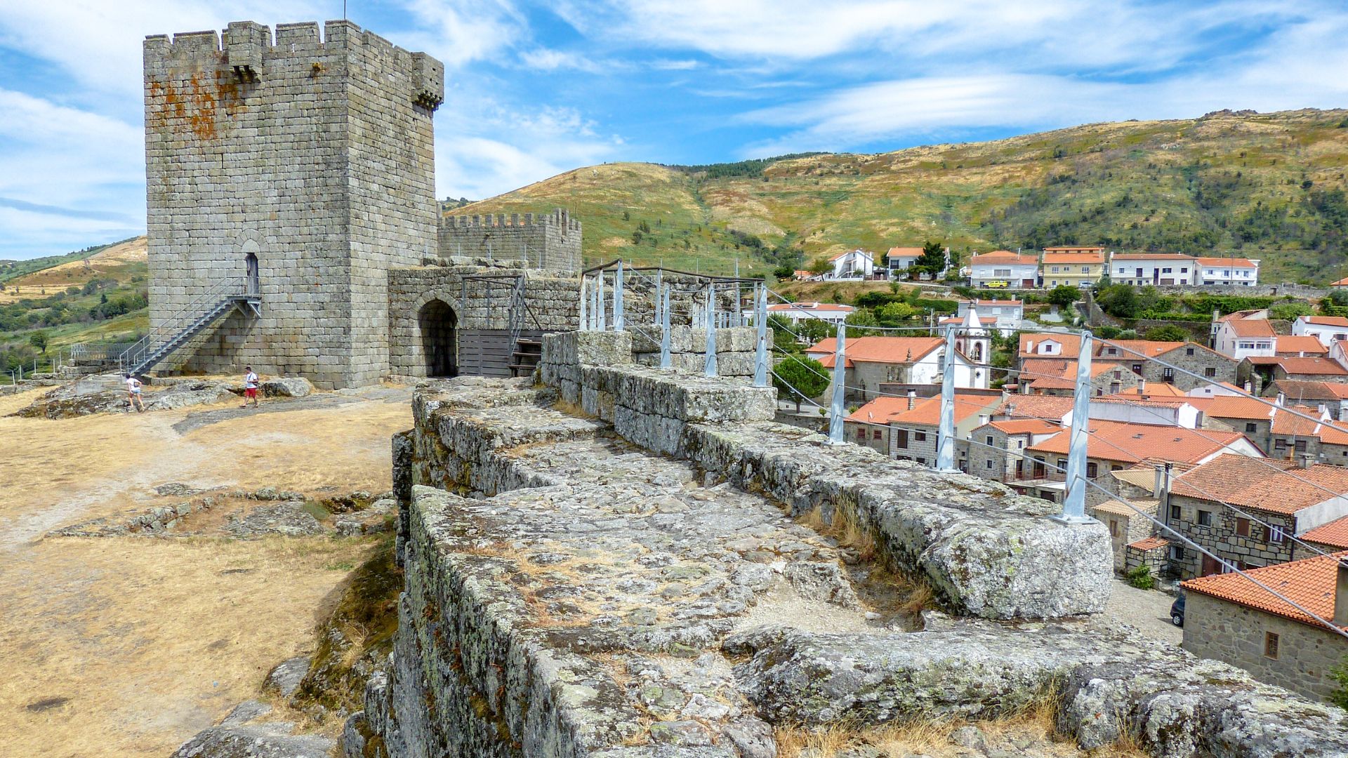 A medieval castle with a prominent keep and fortified walls, situated on a rocky outcrop overlooking a village with terracotta-tiled roofs and a mountainous landscape under a blue sky.