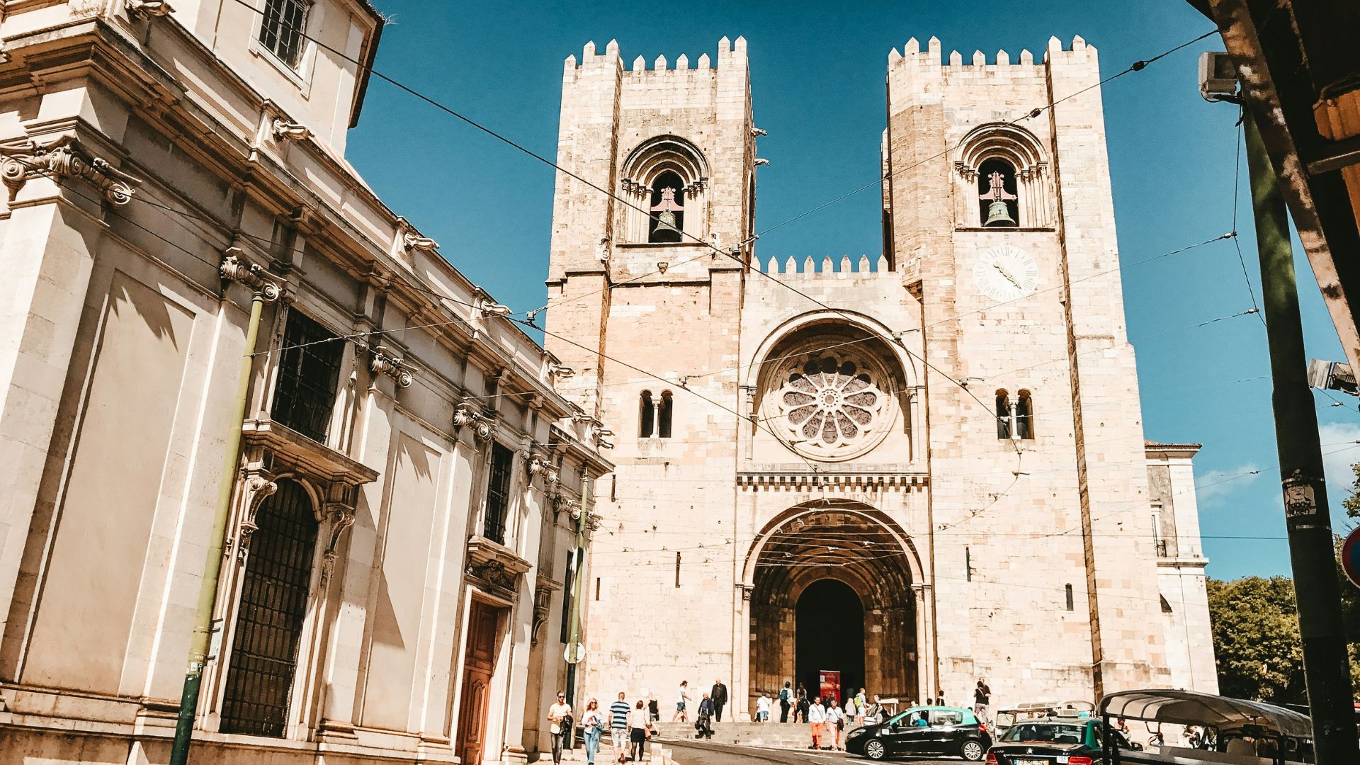 A grand, historic stone cathedral with two prominent bell towers and a large rose window on its facade, situated on a bustling street with people and a car, under a clear blue sky.