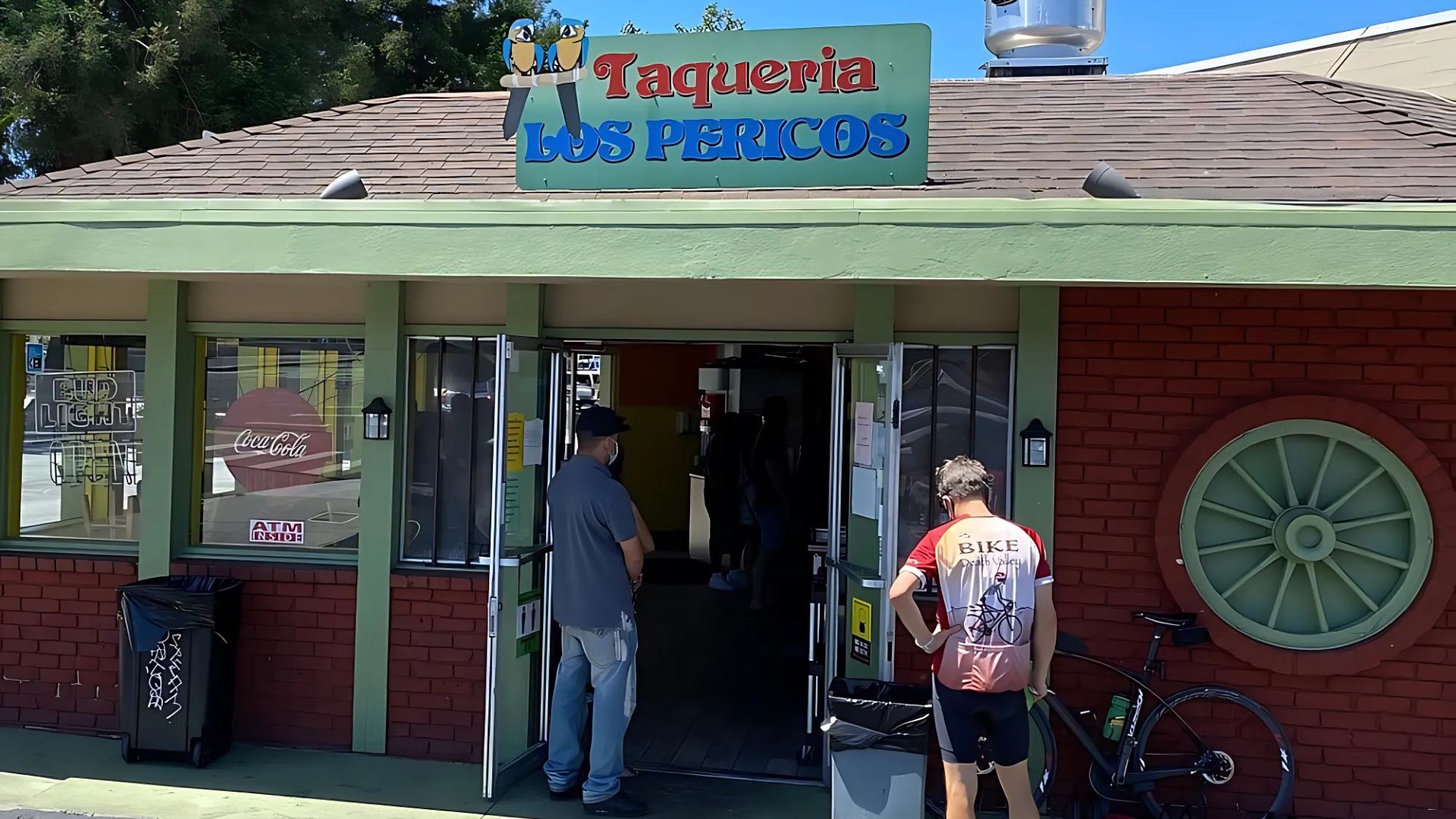 A storefront image of Taqueria Los Pericos, a Mexican restaurant, with two men standing outside the entrance and a bicycle parked on the right. The restaurant sign is prominently displayed above the entrance with "Taqueria LOS PERICOS" written in colorful letters, featuring two parrots at the top. An ATM and Coca-Cola sign are visible through the windows.