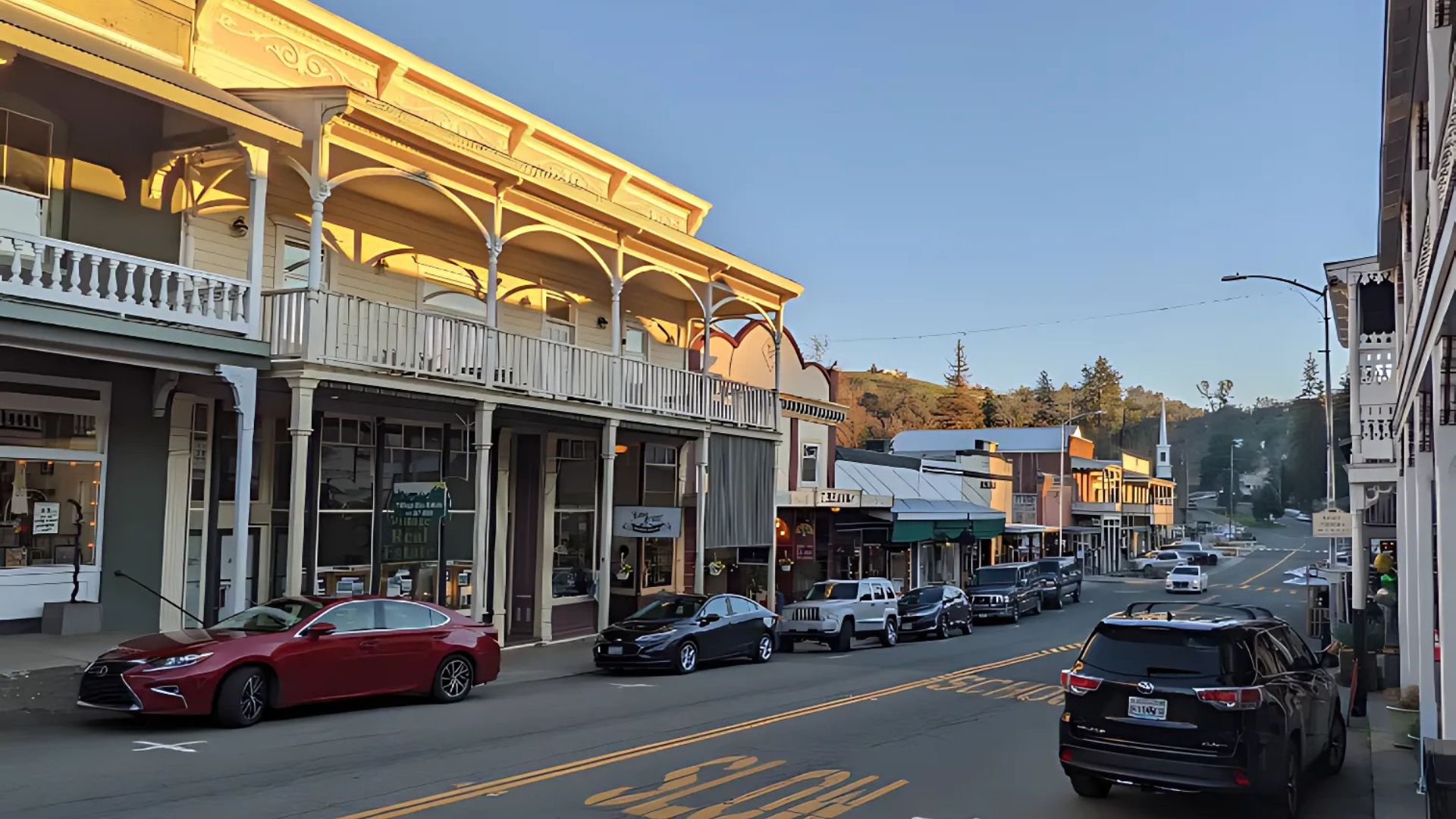 A daytime street scene showing the historic Main Street of Sutter Creek, California, with several cars parked along the right side of the road and a row of ornate, multi-story buildings with balconies lining the left side. The sky is clear, and hills are visible in the background under the golden light of the sun.