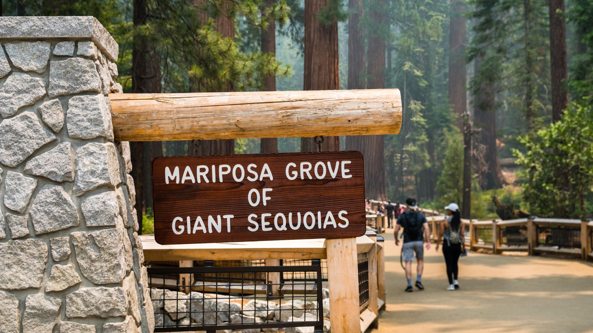 A wooden sign with the text "MARIPOSA GROVE OF GIANT SEQUOIAS" stands next to a stone pillar at the entrance to a forest, with people walking on a path in the background.