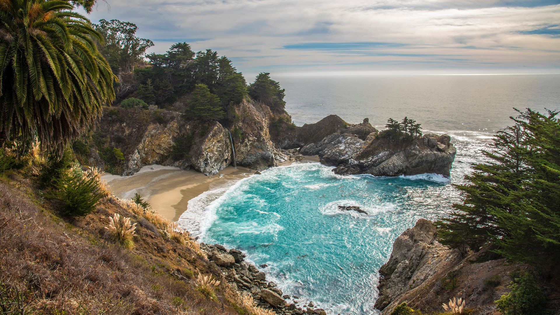 An 80-foot waterfall, McWay Falls, cascading onto a sandy beach within a turquoise cove, surrounded by cliffs and the Pacific Ocean in Big Sur, California.