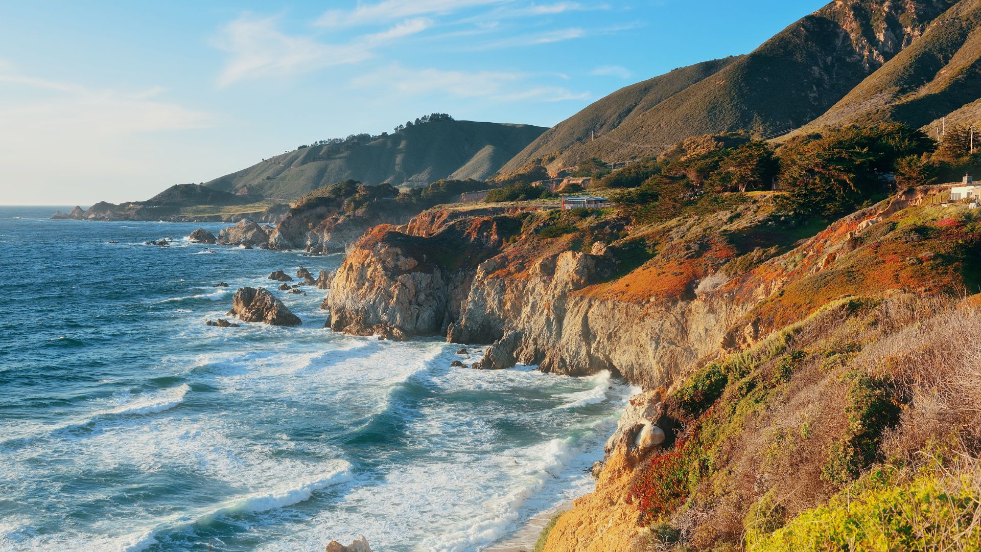 A panoramic view of the rugged Big Sur coastline in California, showing dramatic cliffs meeting the Pacific Ocean with waves crashing against the shore, and verdant hills rising in the background under a clear sky.