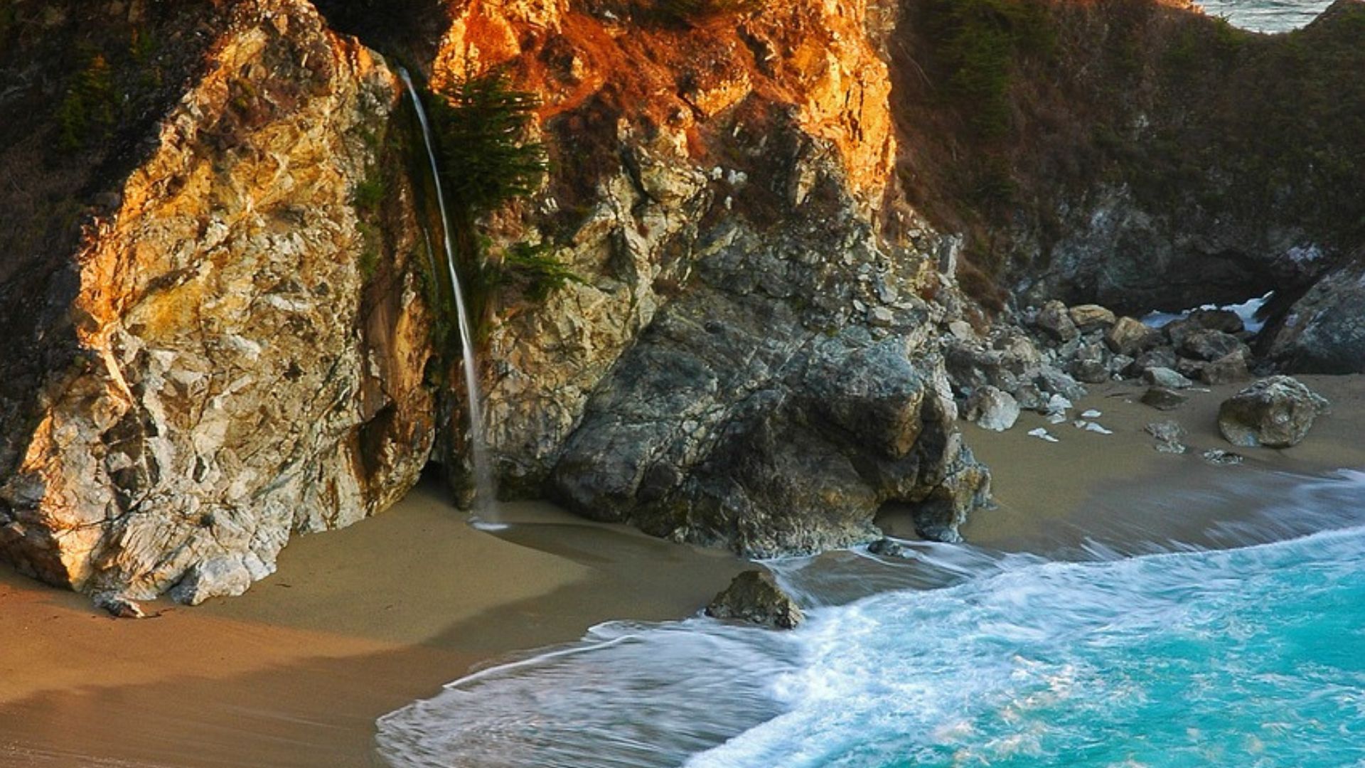 An 80-foot waterfall cascades from a cliff onto a sandy beach within a secluded cove, with turquoise ocean waves breaking on the shore under a golden sunset light, surrounded by lush green trees atop the cliffs.