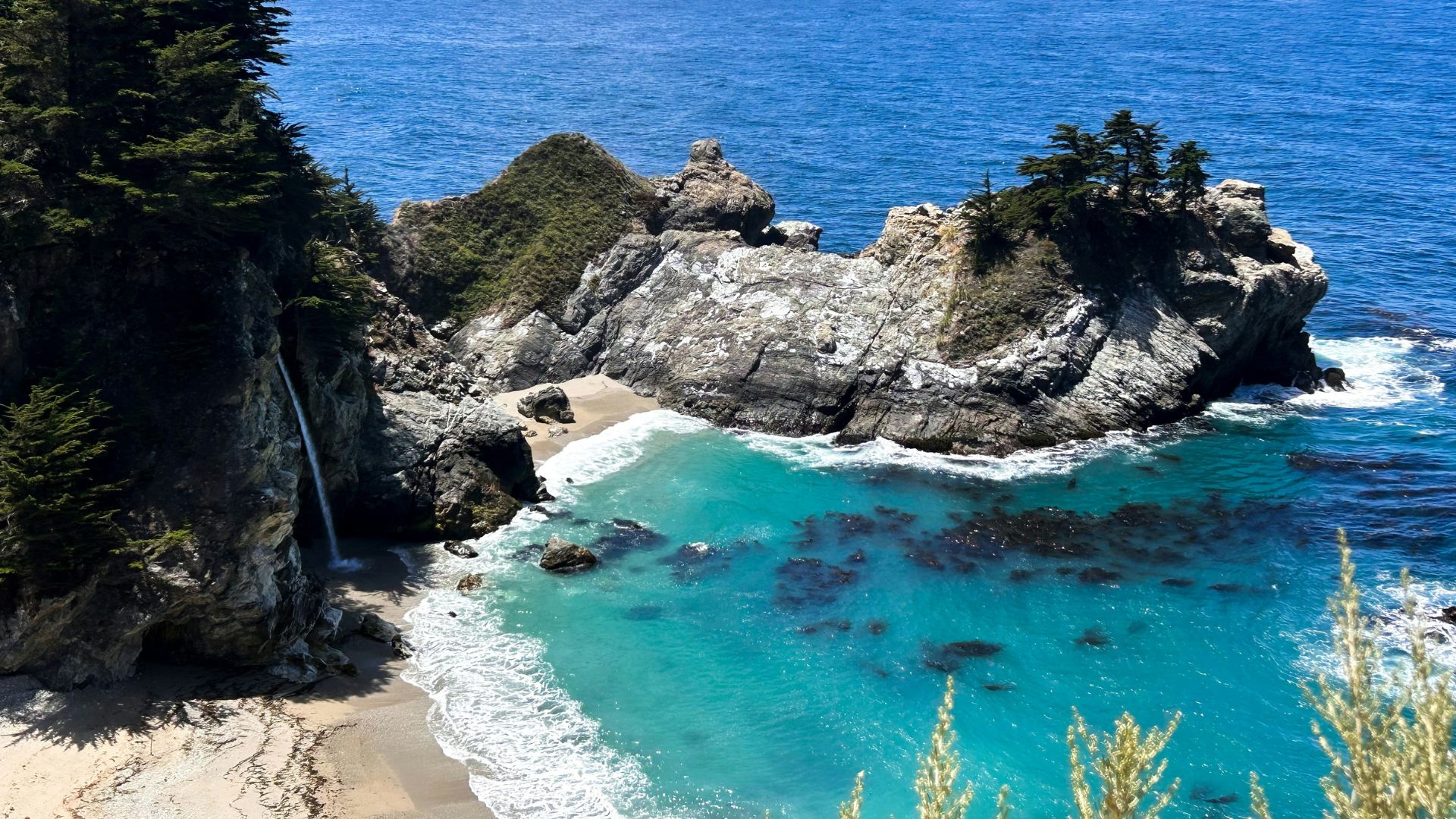 A scenic view of McWay Falls, an 80-foot waterfall cascading onto a sandy cove and into the turquoise waters of the Pacific Ocean, surrounded by rocky cliffs and lush green trees in Big Sur, California.