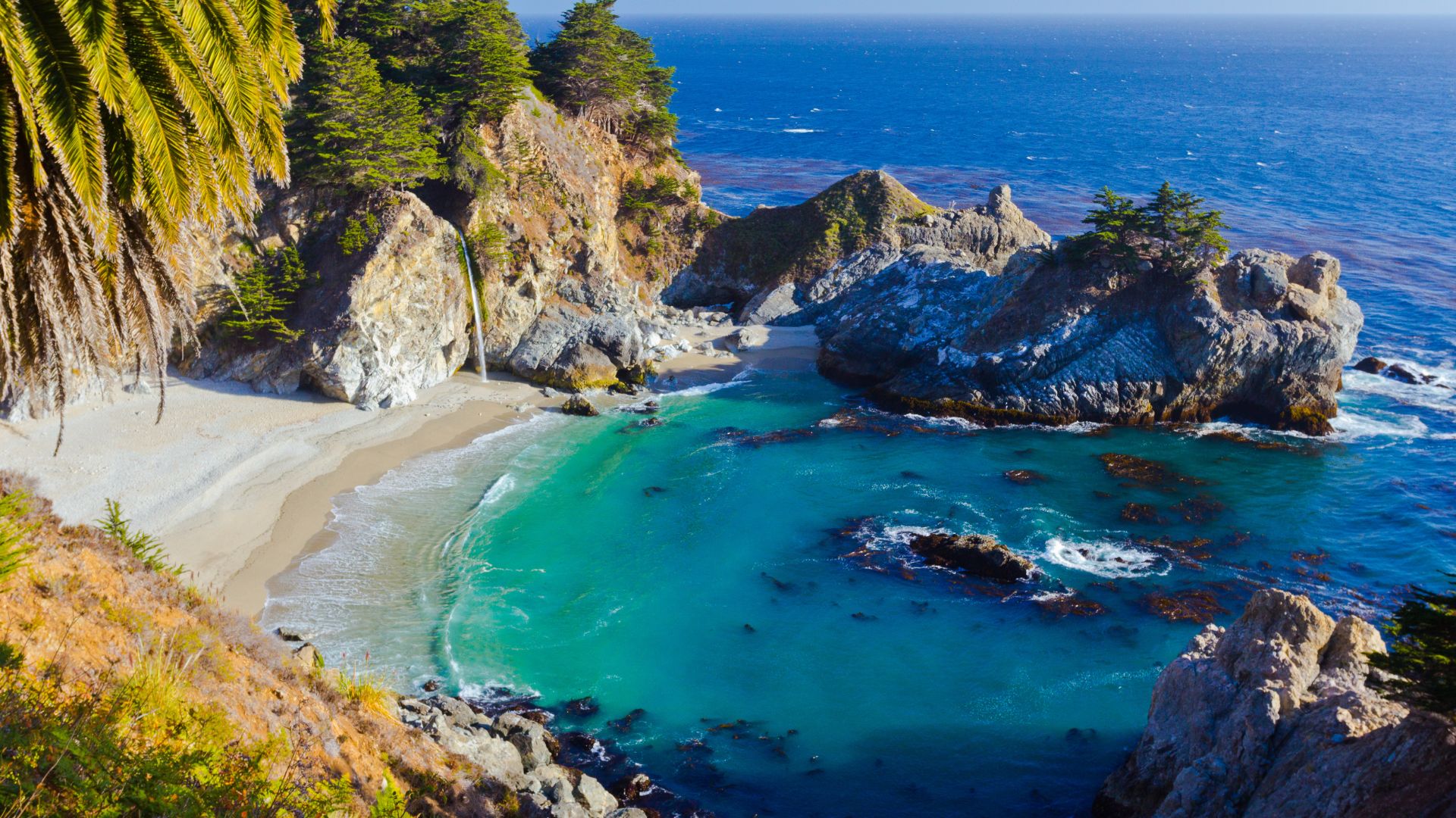 A scenic view of McWay Falls in Big Sur, California, showing an 80-foot waterfall cascading onto a pristine sandy beach within a secluded cove, surrounded by rugged cliffs and lush greenery, with the turquoise Pacific Ocean extending beyond. 
