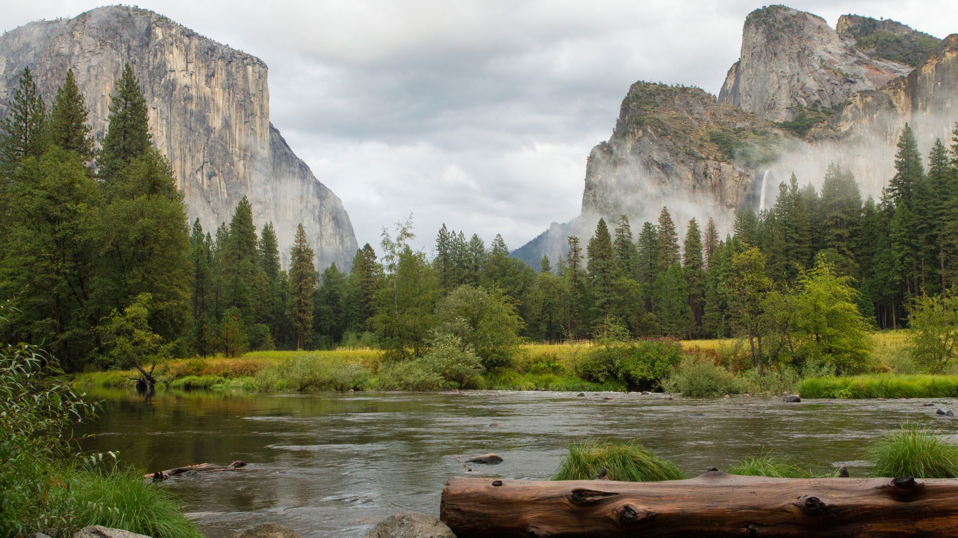 A wide-angle landscape photograph of Yosemite Valley in California, featuring the Merced River flowing in the foreground with a large log partially submerged on the right bank. Towering granite cliffs, including El Capitan on the left and a mist-shrouded mountain with a waterfall (likely Bridalveil Falls) on the right, dominate the background under a cloudy sky. Lush green forests line the riverbanks and extend towards the base of the mountains.
