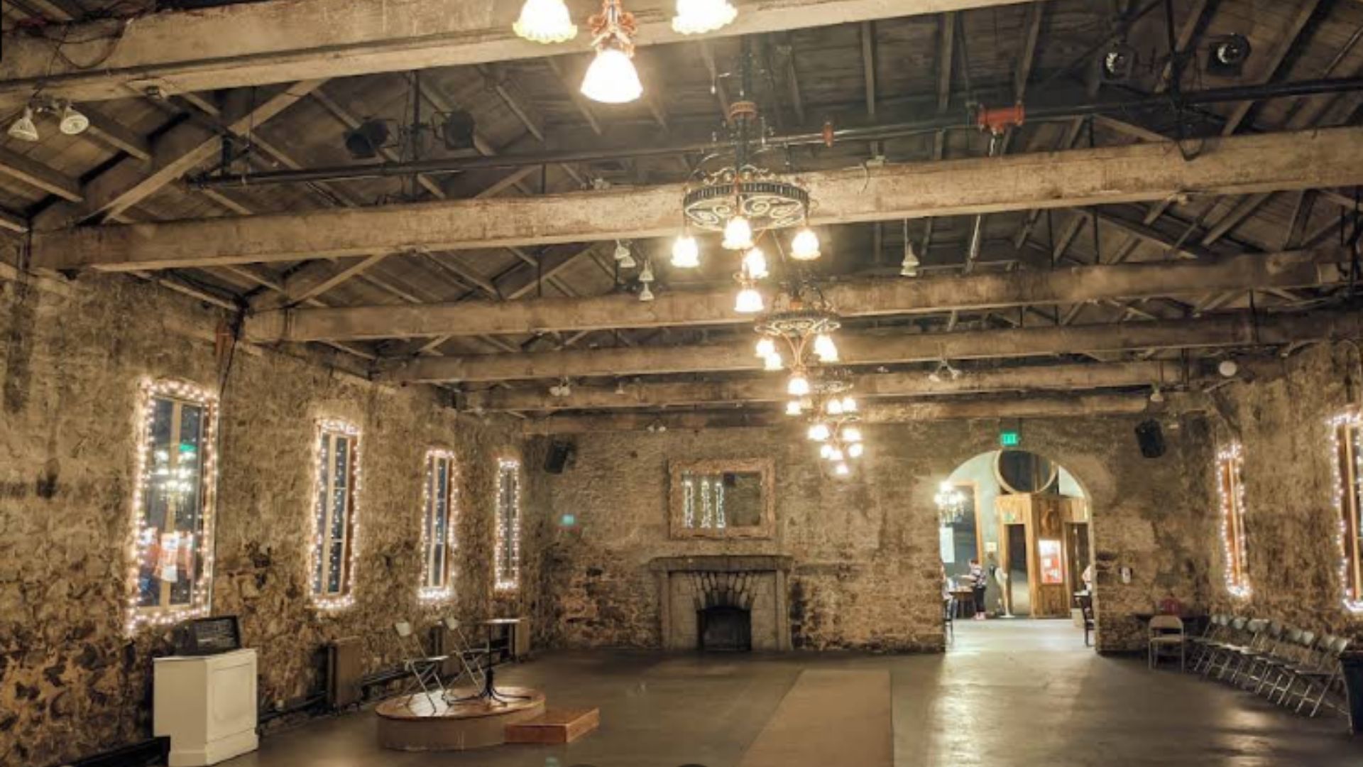 A wide-angle interior view of the Miners Foundry Cultural Center, a large, rustic hall with exposed wooden beams on the ceiling and rough-hewn stone walls. The room is illuminated by multiple chandeliers with exposed bulbs and string lights draped around the windows and a fireplace. Chairs are neatly arranged along the right wall, and a raised platform with a small structure is visible in the foreground on the left. The space appears ready for an event, showcasing its historic charm and architectural features.