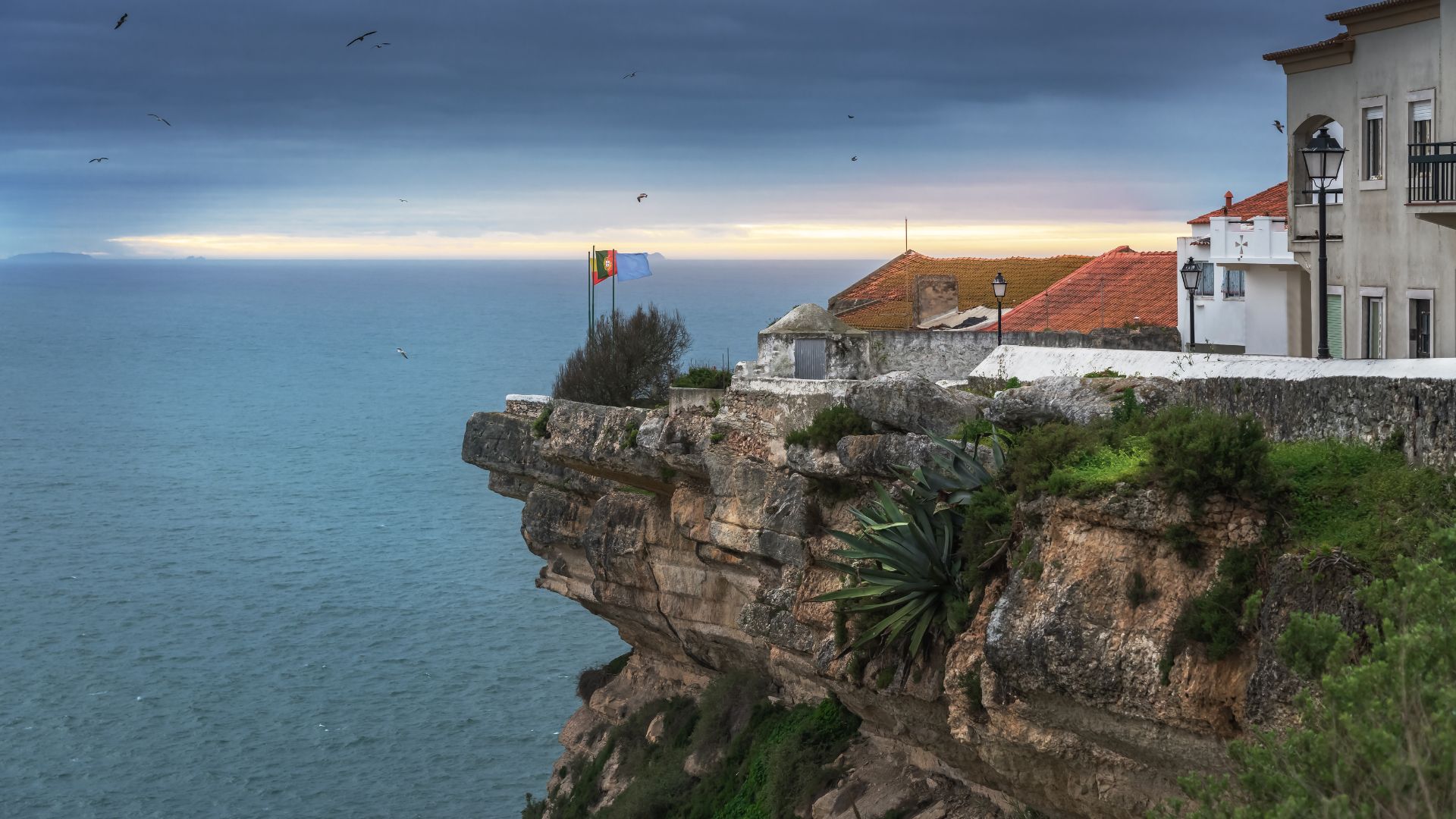 A panoramic view of the Miradouro do Suberco viewpoint in Nazaré, Portugal, featuring a cliffside town with white buildings and red-tiled roofs overlooking the vast, cloudy Atlantic Ocean under a dramatic sky, with a flag flying near the cliff edge.