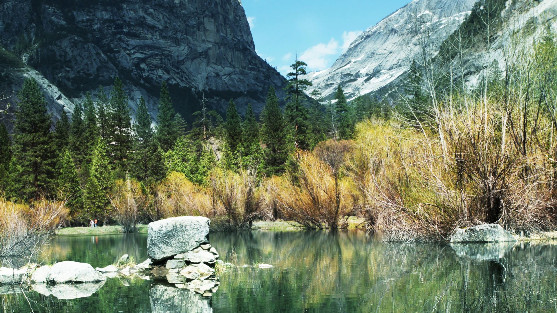 A serene landscape featuring Mirror Lake in Yosemite National Park, with its calm waters reflecting the surrounding towering granite cliffs and lush green and golden-hued trees. Large boulders are visible in the foreground of the lake, under a clear blue sky. 