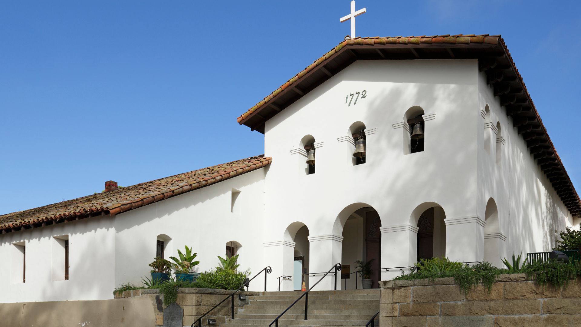 A white, adobe-style mission building with a red tile roof and a cross on its facade, featuring arched entrances and bell openings, with the year "1772" visible, set against a clear blue sky. 
