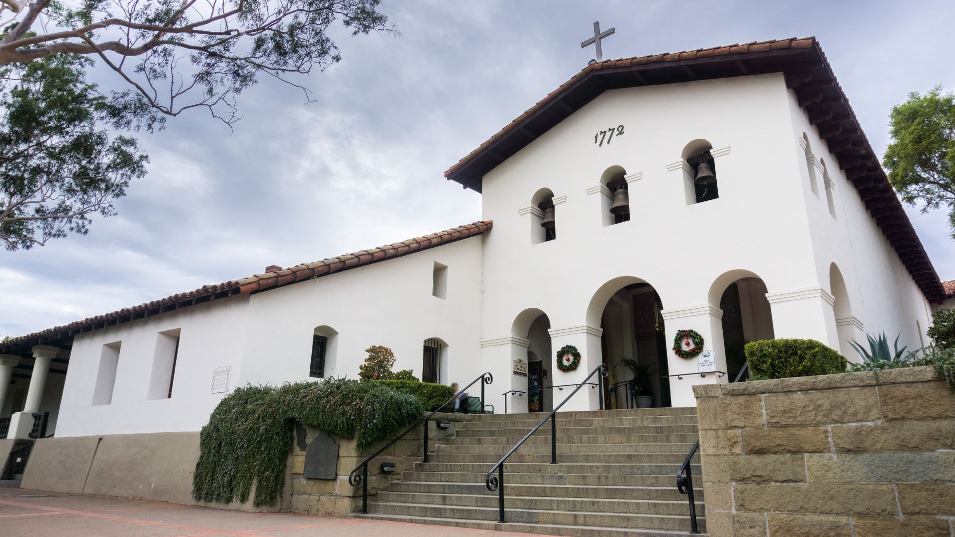 A low-angle shot of the Mission San Luis Obispo de Tolosa, a white Spanish colonial-style church with a tiled roof and bell arches, featuring a prominent cross atop its facade and the year "1772" inscribed below the bells. Stone steps lead up to the arched entrance, flanked by lush green foliage and wreaths on the doors, all set against a cloudy sky. 