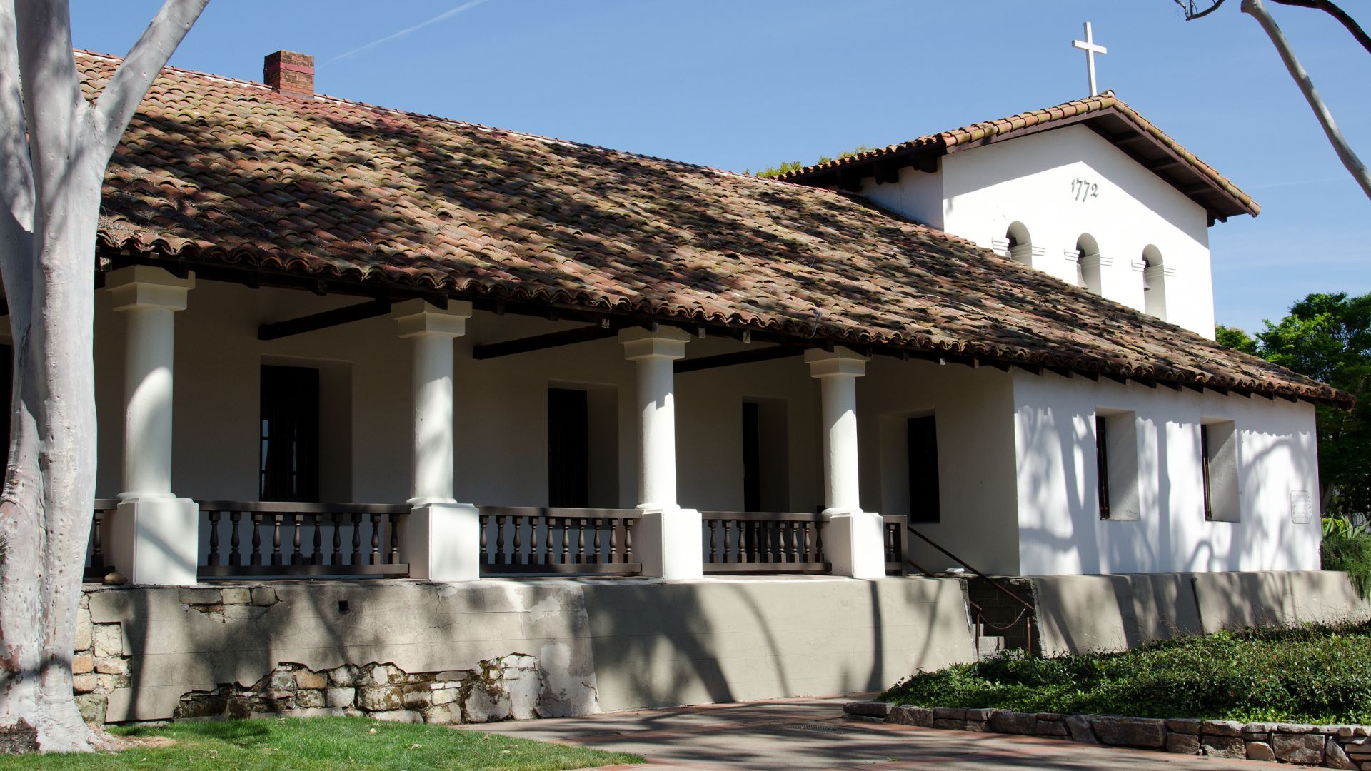 A historic Spanish mission building, Mission San Luis Obispo de Tolosa, features a white facade with a colonnaded porch, a brown tiled roof, and a bell tower topped with a cross on the right side of the structure. The mission is set against a clear blue sky, with a large tree on the left and green foliage in the foreground. 
